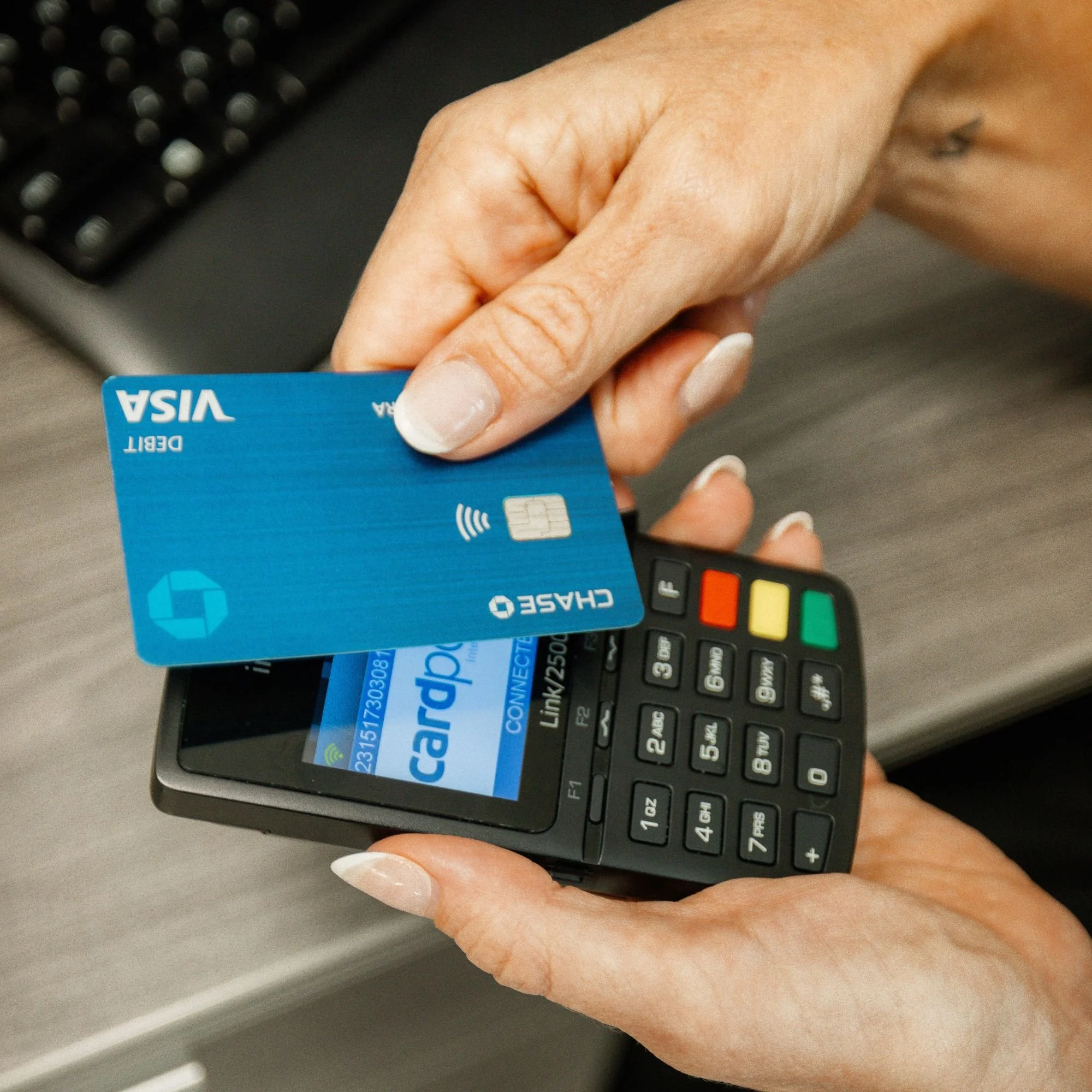 A person holding a blue Visa debit card over a black remote control with color-coded buttons, with a computer keyboard in the background.