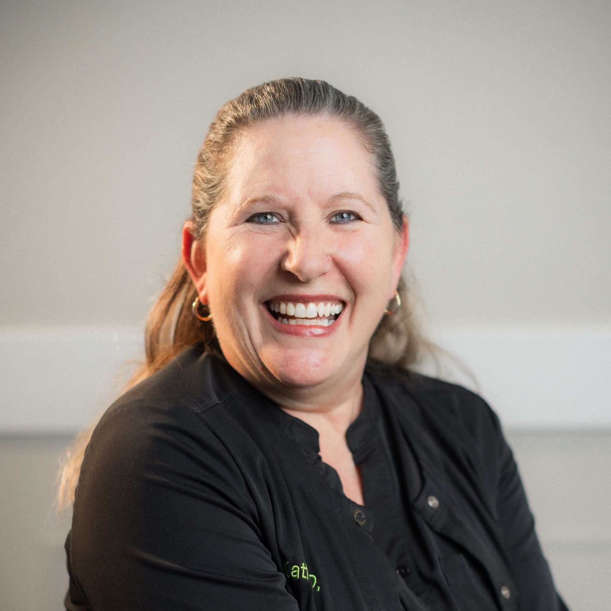 A smiling woman with light skin, brown hair, and blue eyes wearing a black top, with a light neutral background.