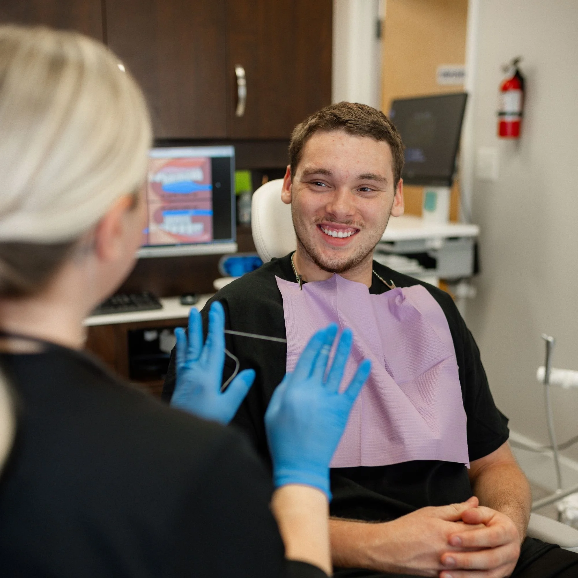 A young man with brown hair and a beard smiling as he interacts with a healthcare worker who is wearing blue gloves and a black shirt. The healthcare worker's hand is visible in the foreground, and the scene appears to be taking place in a clinical or medical setting with computers and a fire extinguisher for safety.