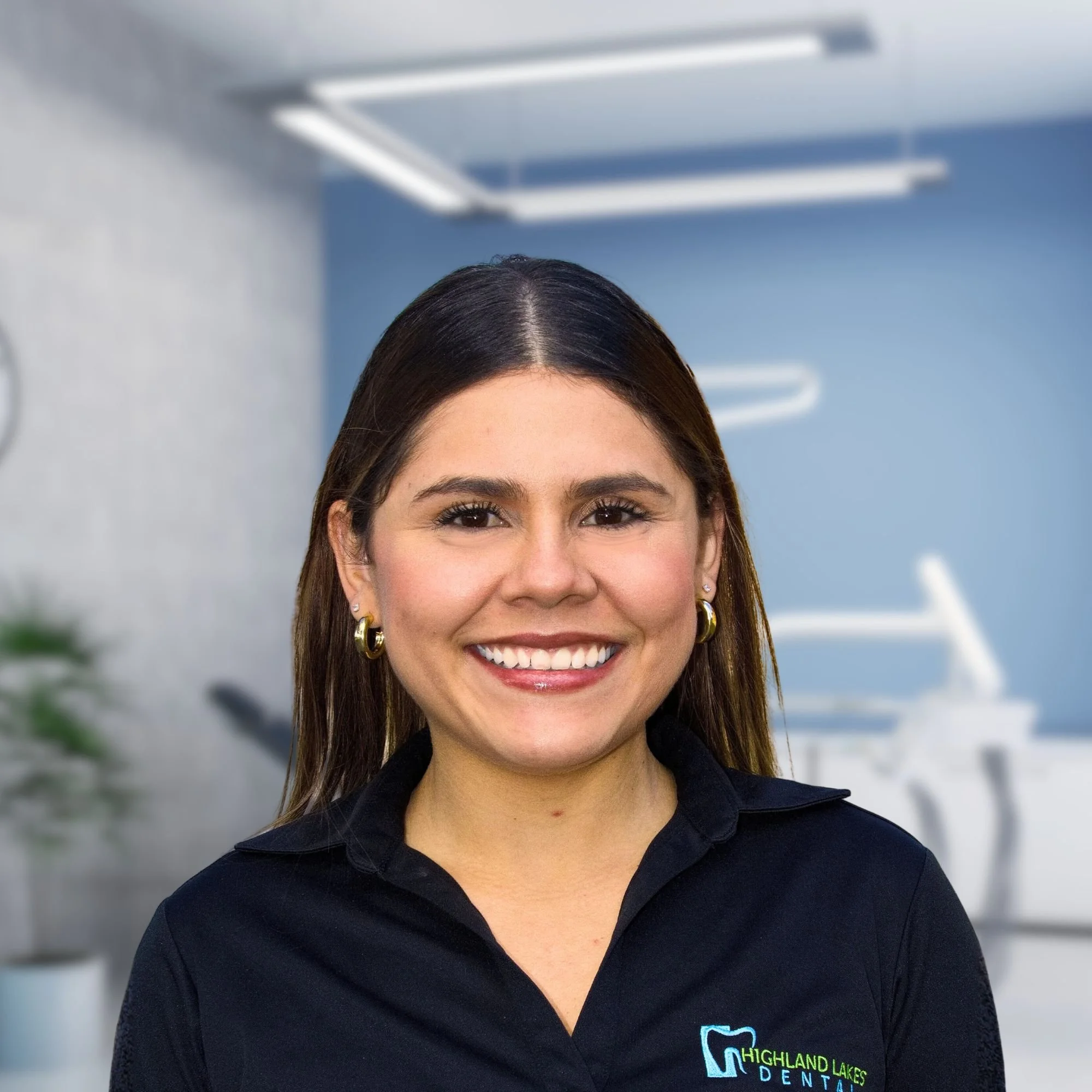 Close-up of a smiling woman wearing a black Highland Lakes Dental polo shirt, with brown hair and earrings, standing indoors with blue walls and fluorescent lighting.