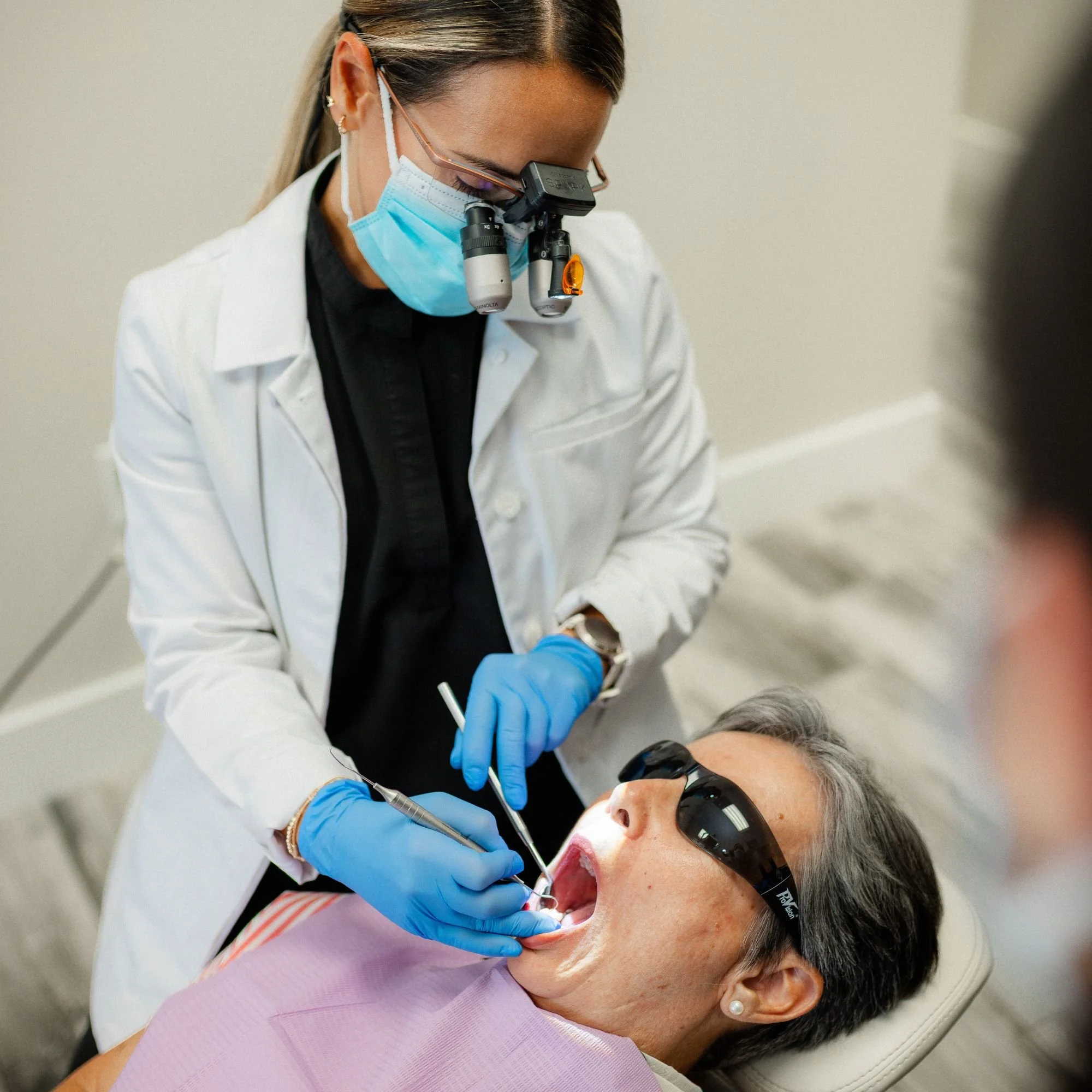 Dentist wearing blue gloves and a face mask performing a dental procedure on a patient lying back in the dental chair with protective glasses.