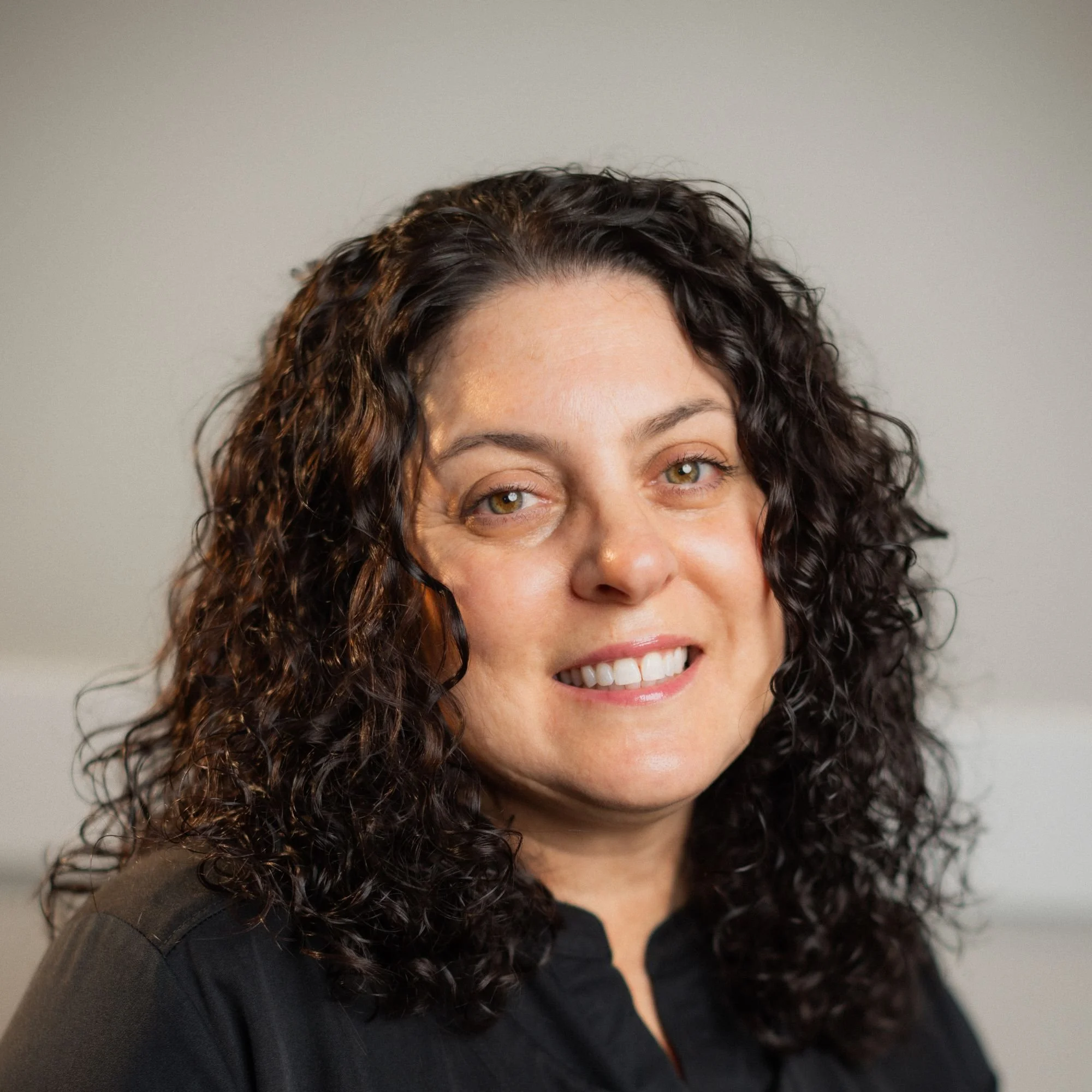 Close-up of a woman with curly dark hair, smiling with visible teeth, wearing a black top, against a plain grey background.