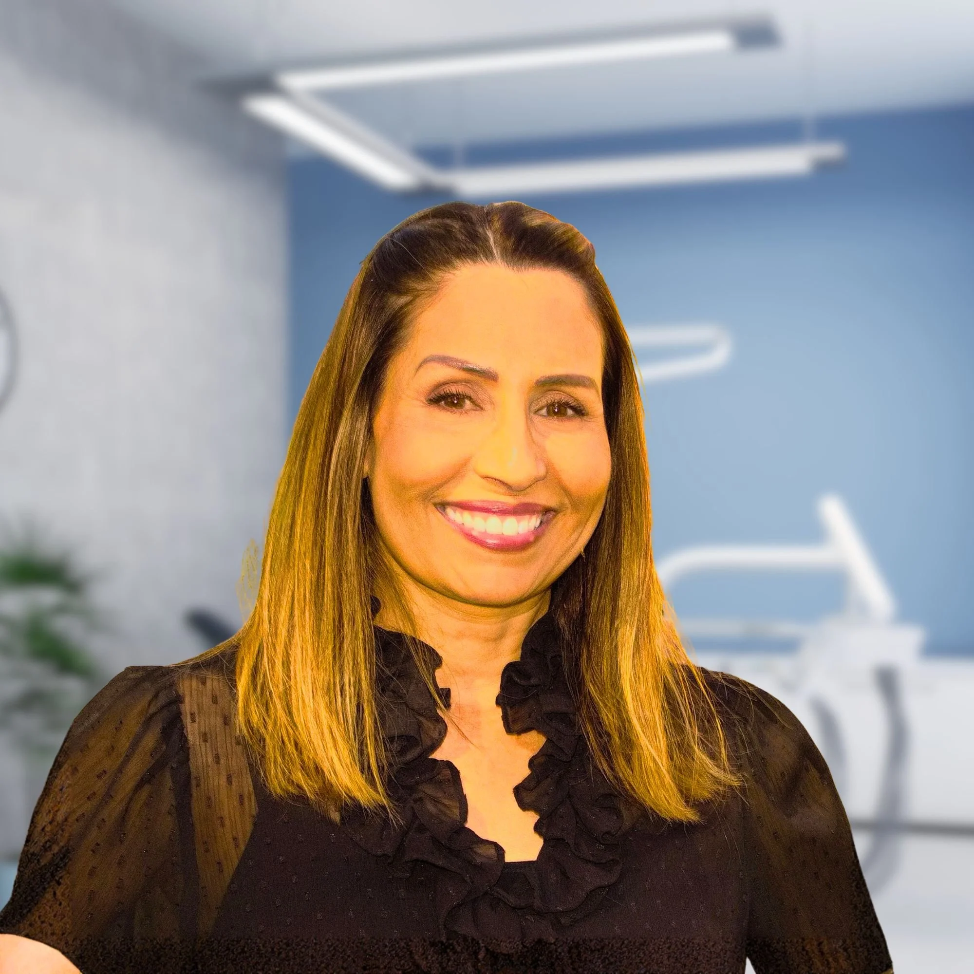 Close-up of a woman with long brown hair and smiling face, wearing a black sheer lace top, against a blue background with blurred ceiling lights.