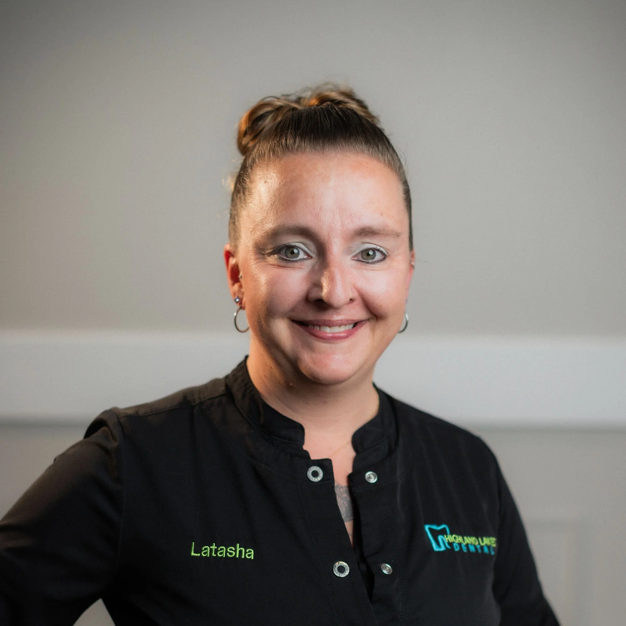A woman with short, dark hair styled in a bun, wearing a black shirt with green text that says "Latasha" and a logo that reads "Highland Lakes Dental," taking a selfie against a gray background.