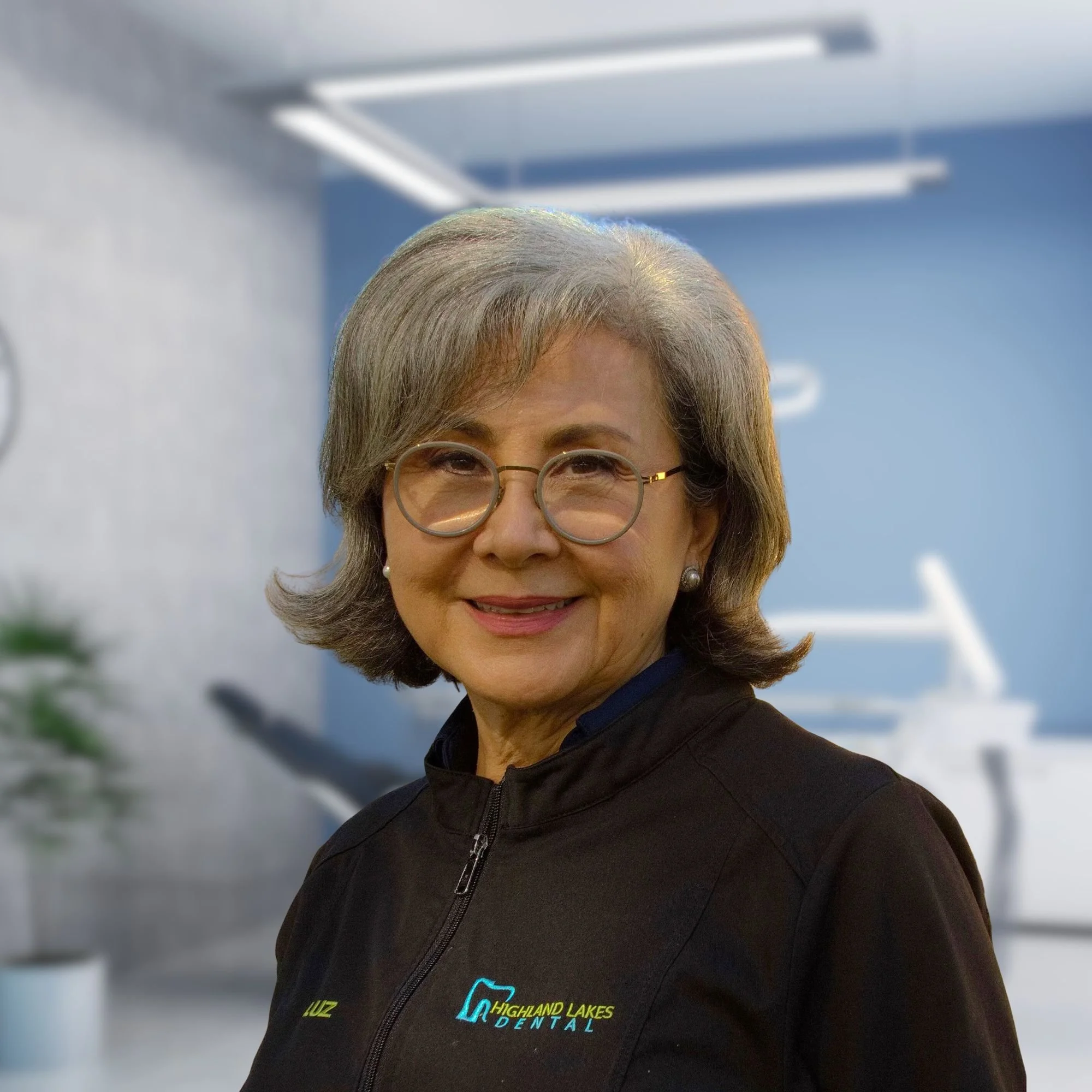 Close-up of a woman wearing glasses and a black jacket with the Highland Lakes Dental logo, standing outdoors with snow and blue sky in the background.
