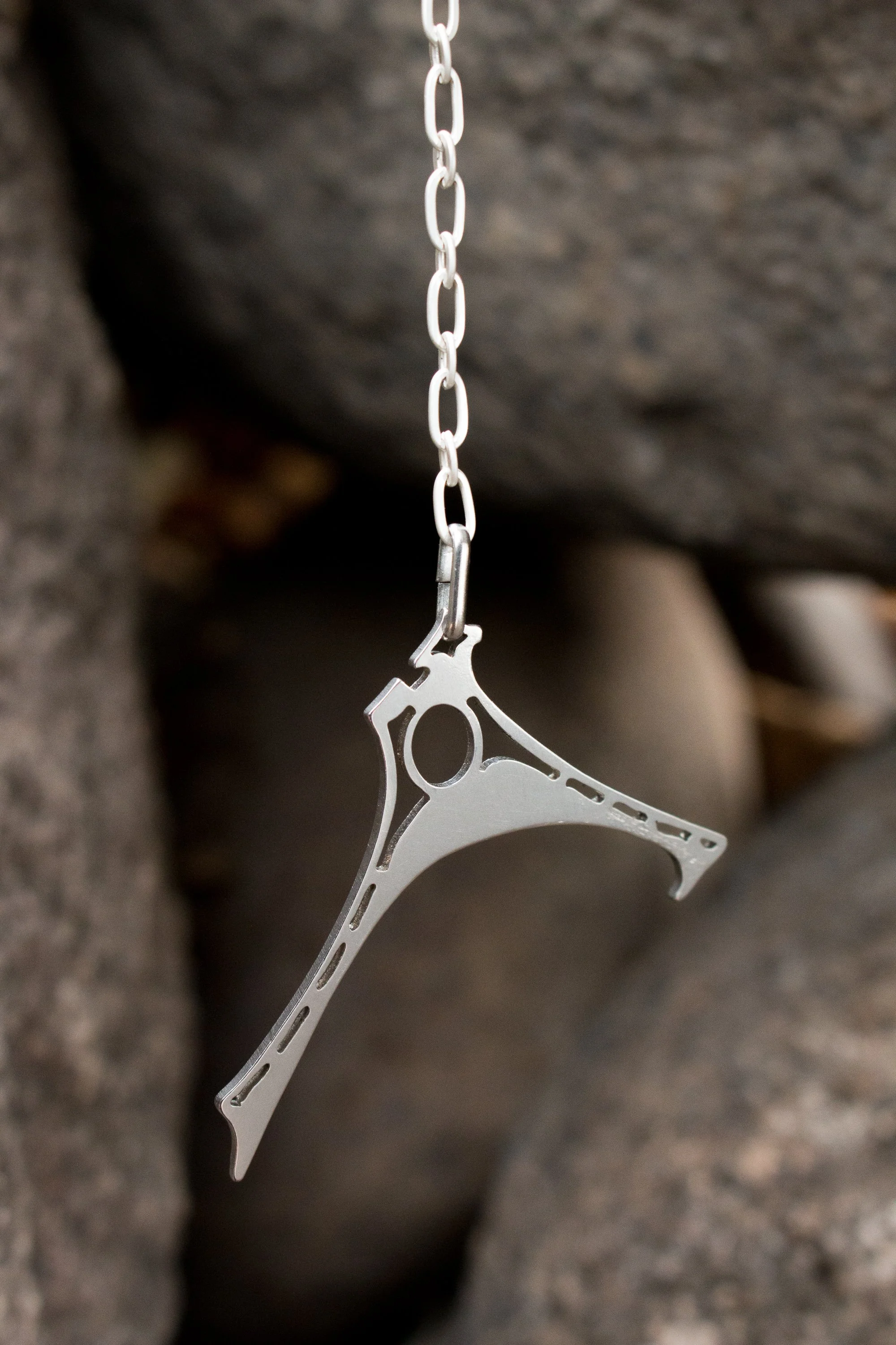 The pendant hanging from a silver chain against soft-focus stone, showing the asymmetric curved form and circular cut-out.