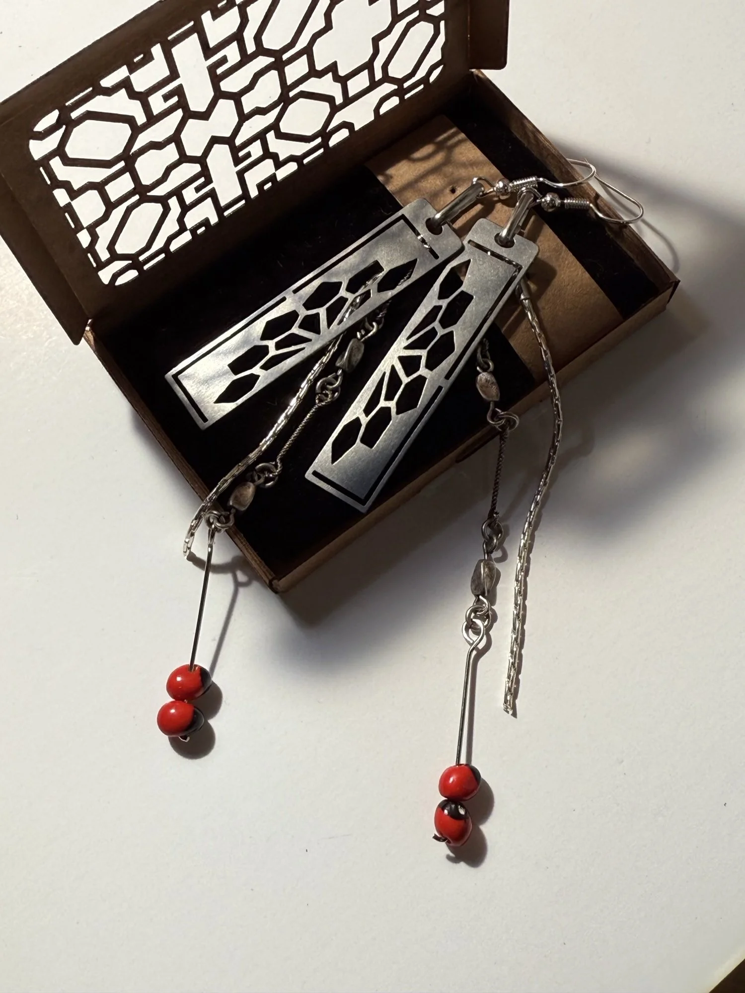 The earrings resting in a box with a cut-out lid, red beads trailing onto the surface below. (Copy)