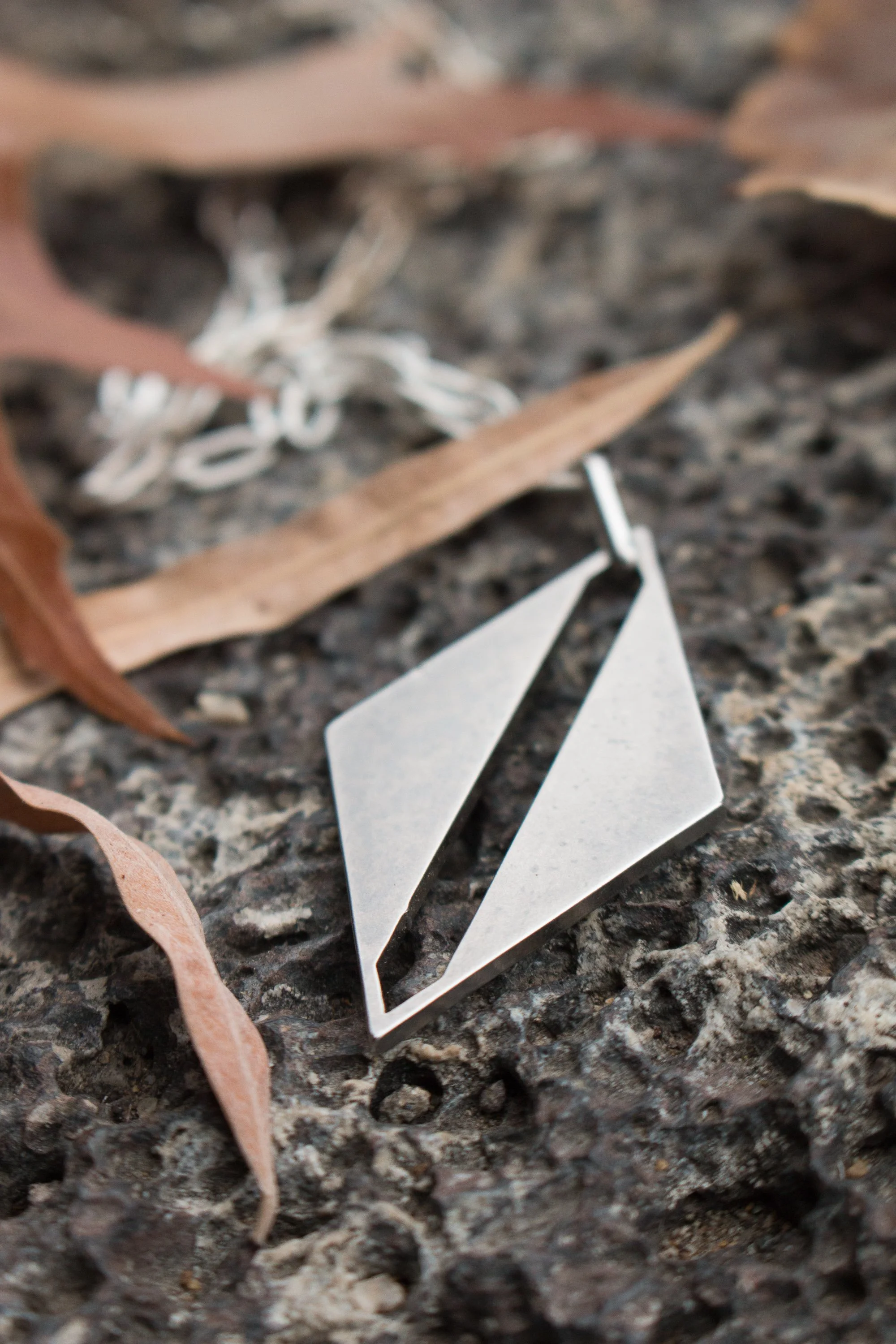 Hand-fabricated stainless steel diamond pendant with asymmetric cut-out detail, resting on weathered stone beside dry leaves.