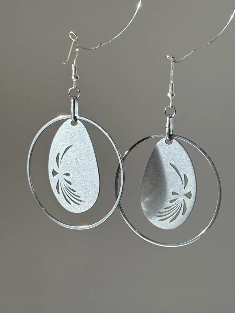 Hand-fabricated stainless steel hoop earrings with oval medallion and radiant cut-out detail, hanging against a gray background.