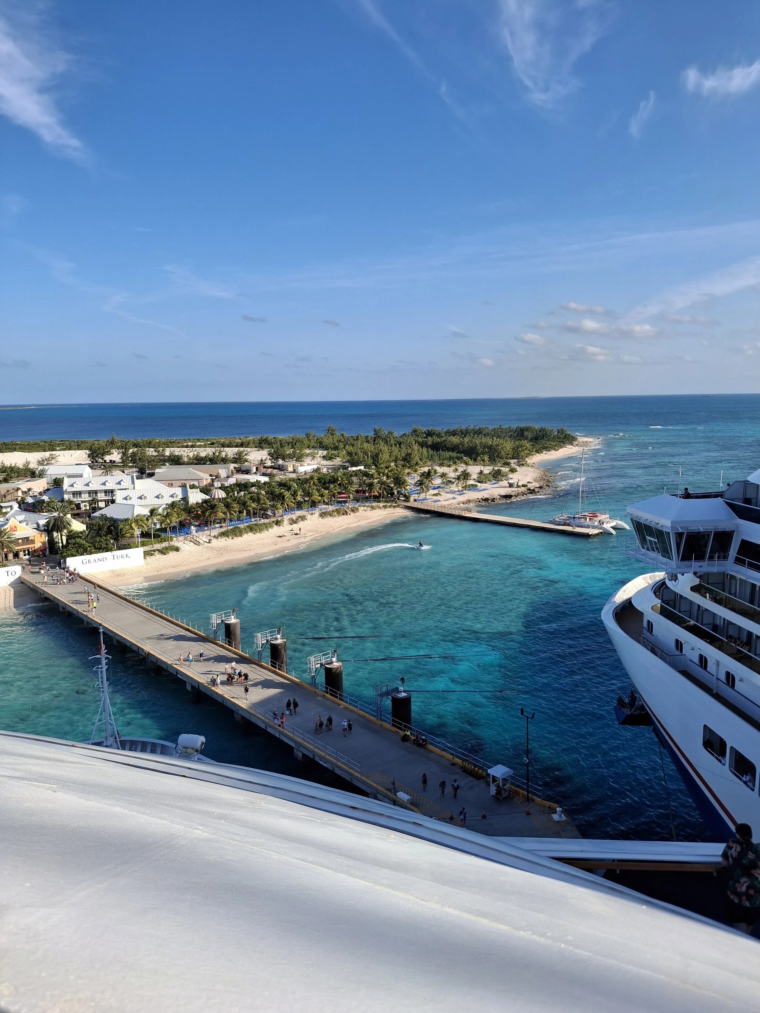 Aerial view of a cruise ship docked at a tropical island port, with turquoise waters, a pier with people, green trees, a sandy beach, and a clear blue sky with some clouds.