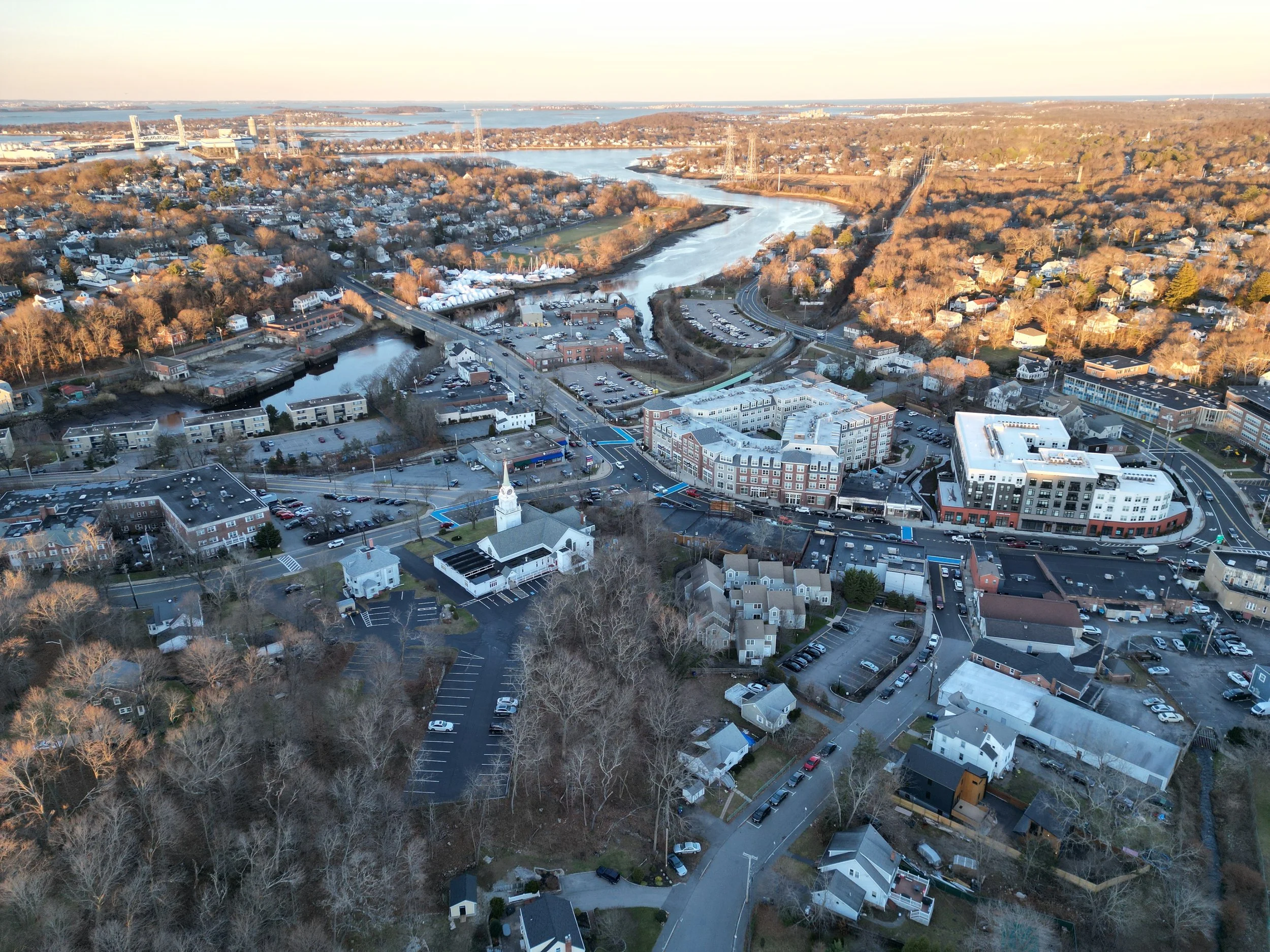 Aerial view of a city with a river running through it, surrounded by residential and commercial buildings, with bridges and a distant water body.