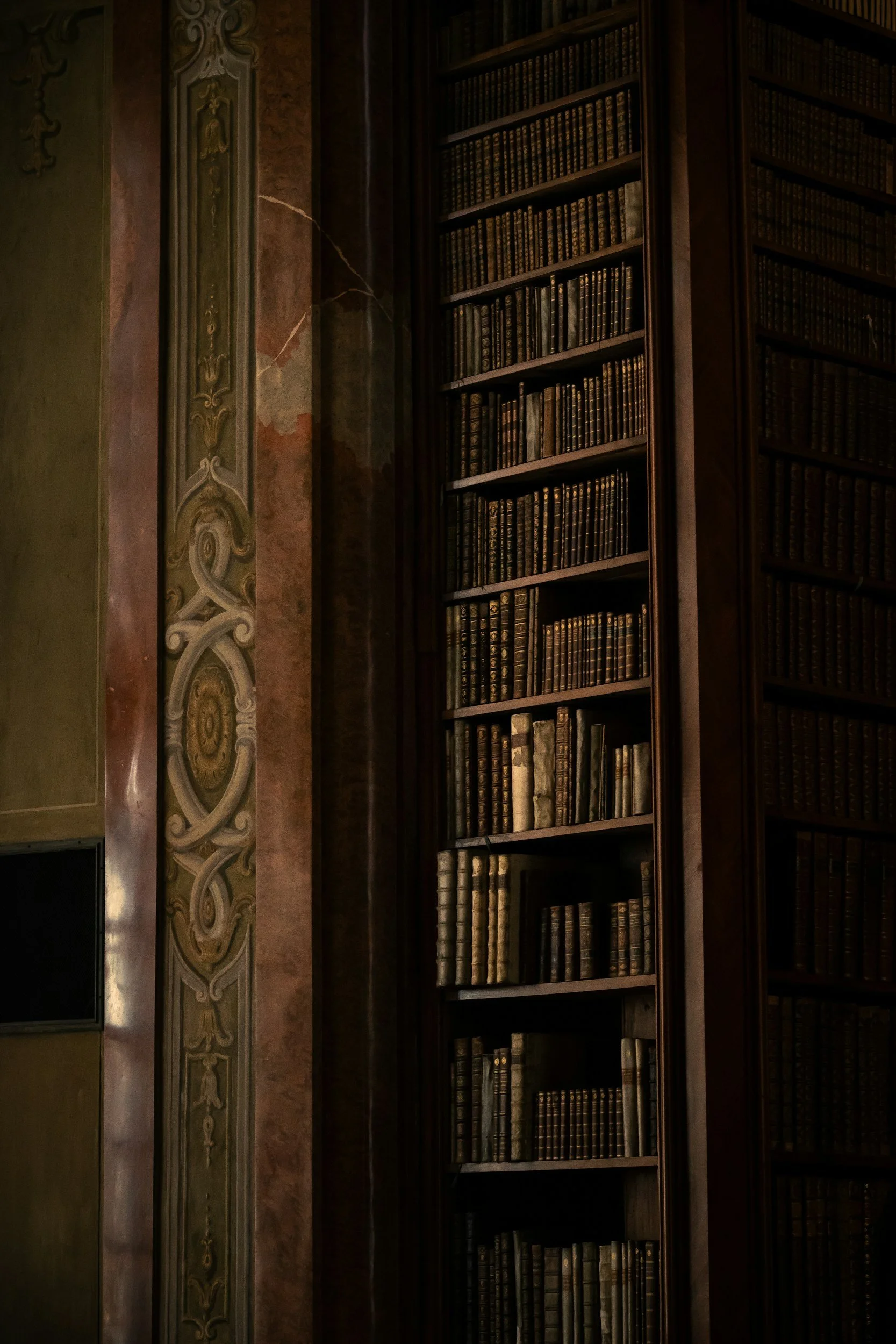 A tall wooden bookshelf filled with old, leather-bound books next to a decorative painted wall in a historic library or study.