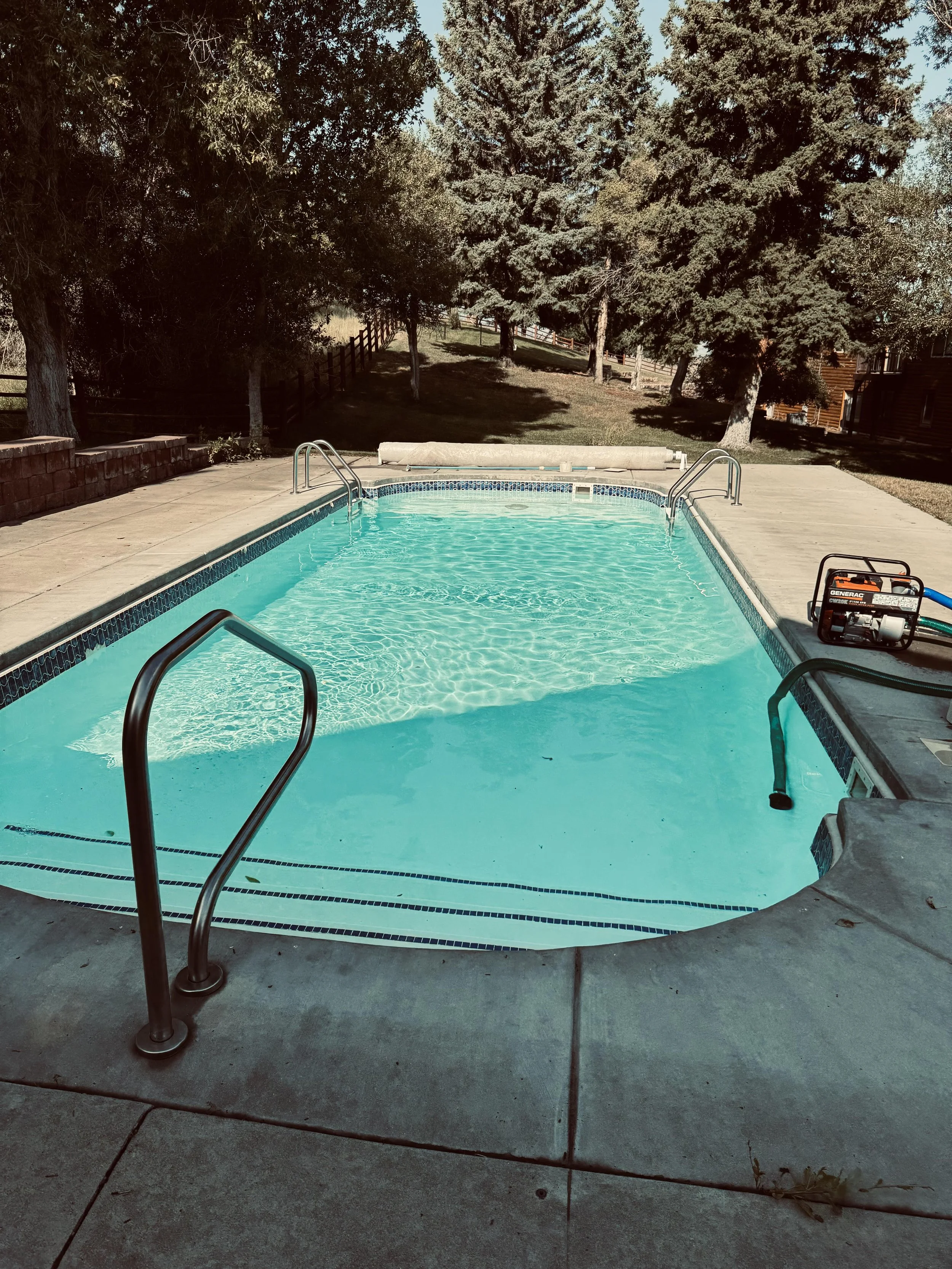An outdoor swimming pool with clear water surrounded by a concrete deck, with a ladder on the left side and a pool cleaner on the right side, set in a backyard with trees and a grassy hill in the background.