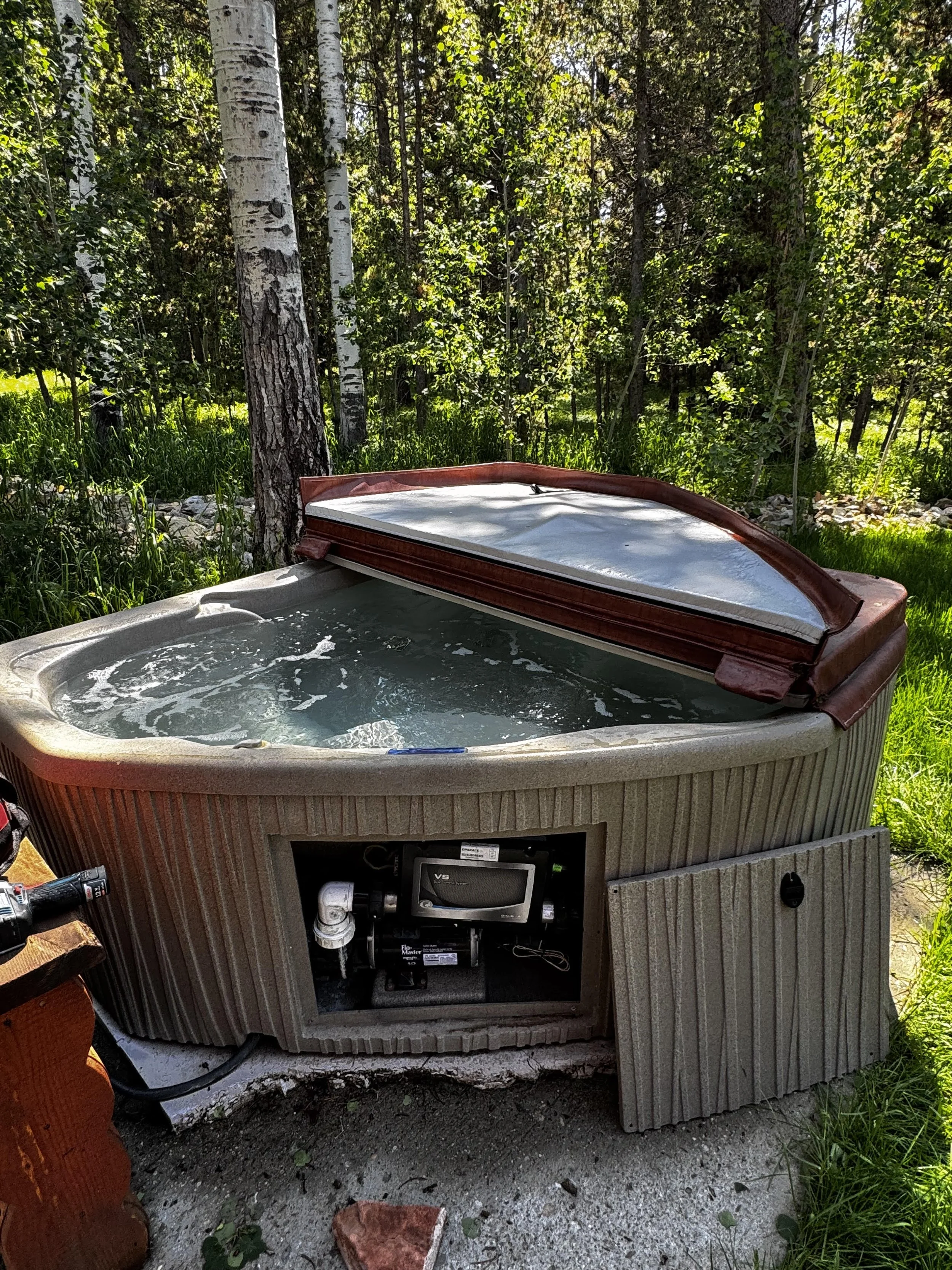 Hot tub with cover open, centered in an outdoor area with grass and trees; part of the hot tub equipment visible underneath.