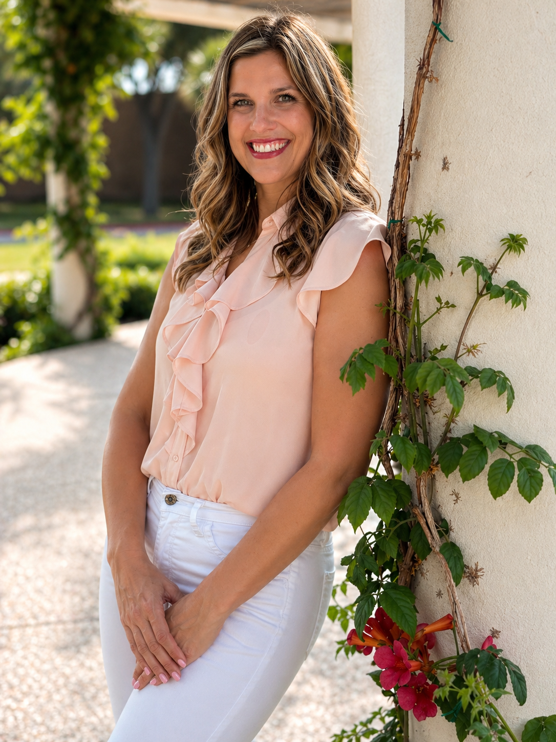 Smiling woman with wavy blonde hair wearing a peach blouse with ruffles and white pants, leaning against a vine-covered wall outdoors.
