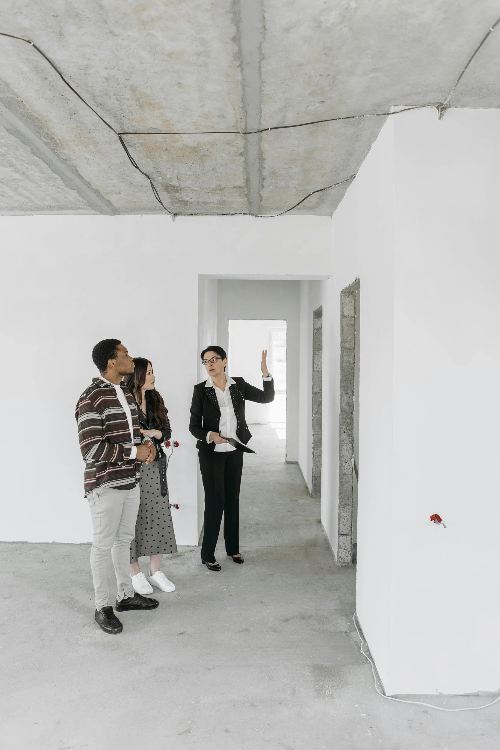 Group of four people, three women and one man, standing in an unfinished room with concrete floor and walls. The woman on the right, wearing glasses and a black suit, appears to be explaining something using gestures, while the other three listen.