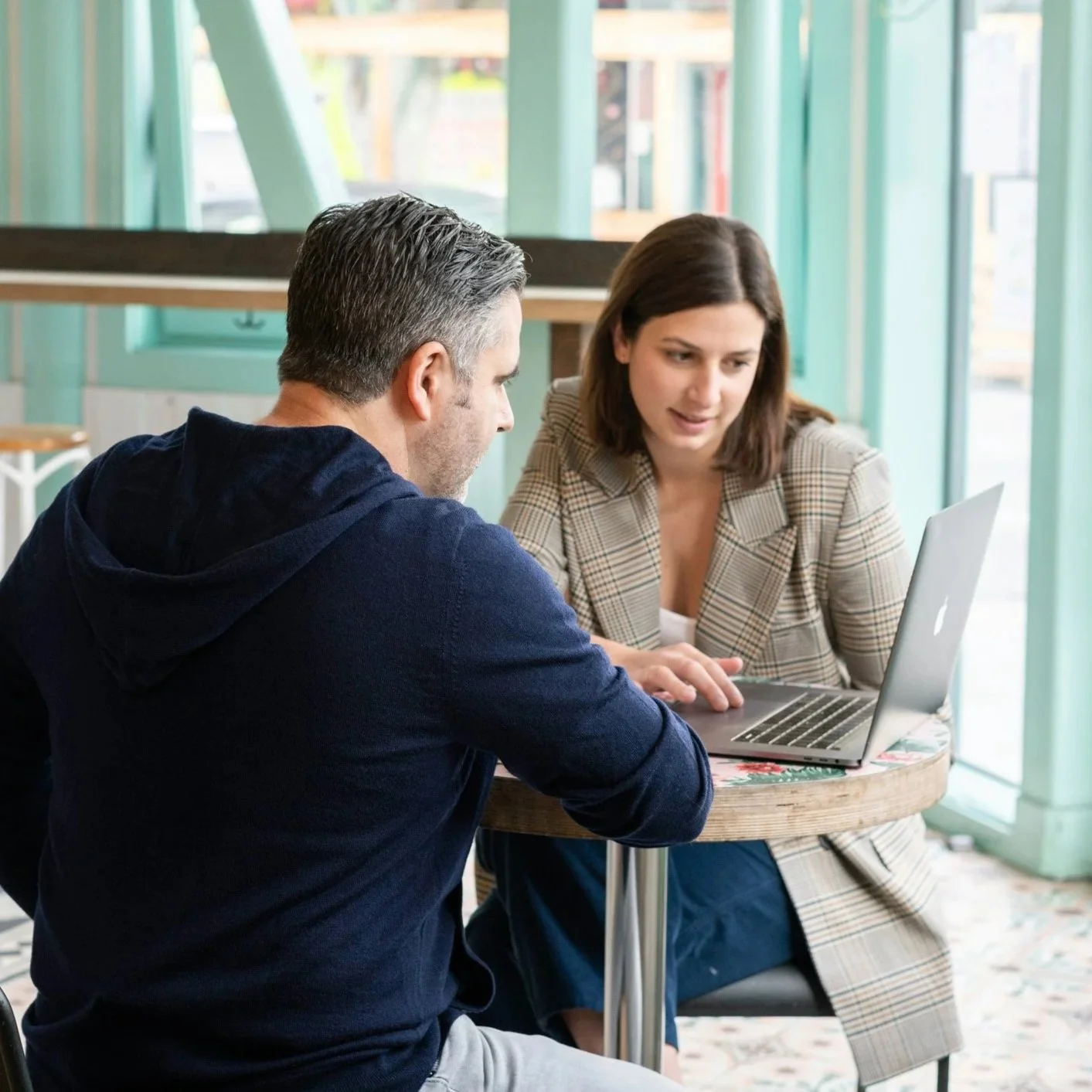 A man with dark hair wearing a dark hoodie and a woman with brown hair wearing a plaid blazer are sitting at a small round table with a laptop, engaging in a discussion in a brightly lit cafe with green frames and large windows.