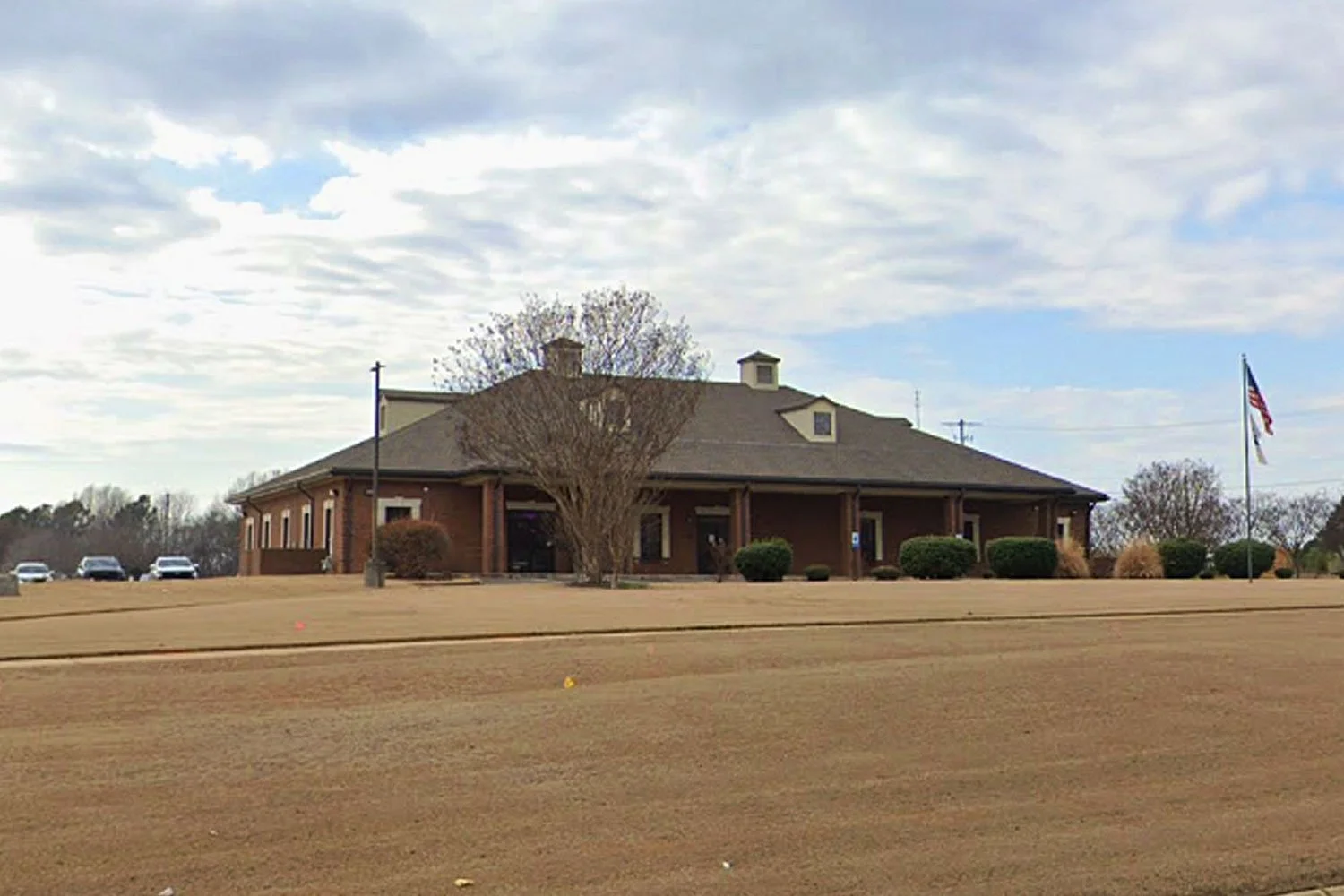 A single-story brick building with a pitched roof and small chimney, surrounded by a lawn with shrubs and a large leafless tree in front, two American flags on flagpoles, and a parking lot with a few cars, under a partly cloudy sky.