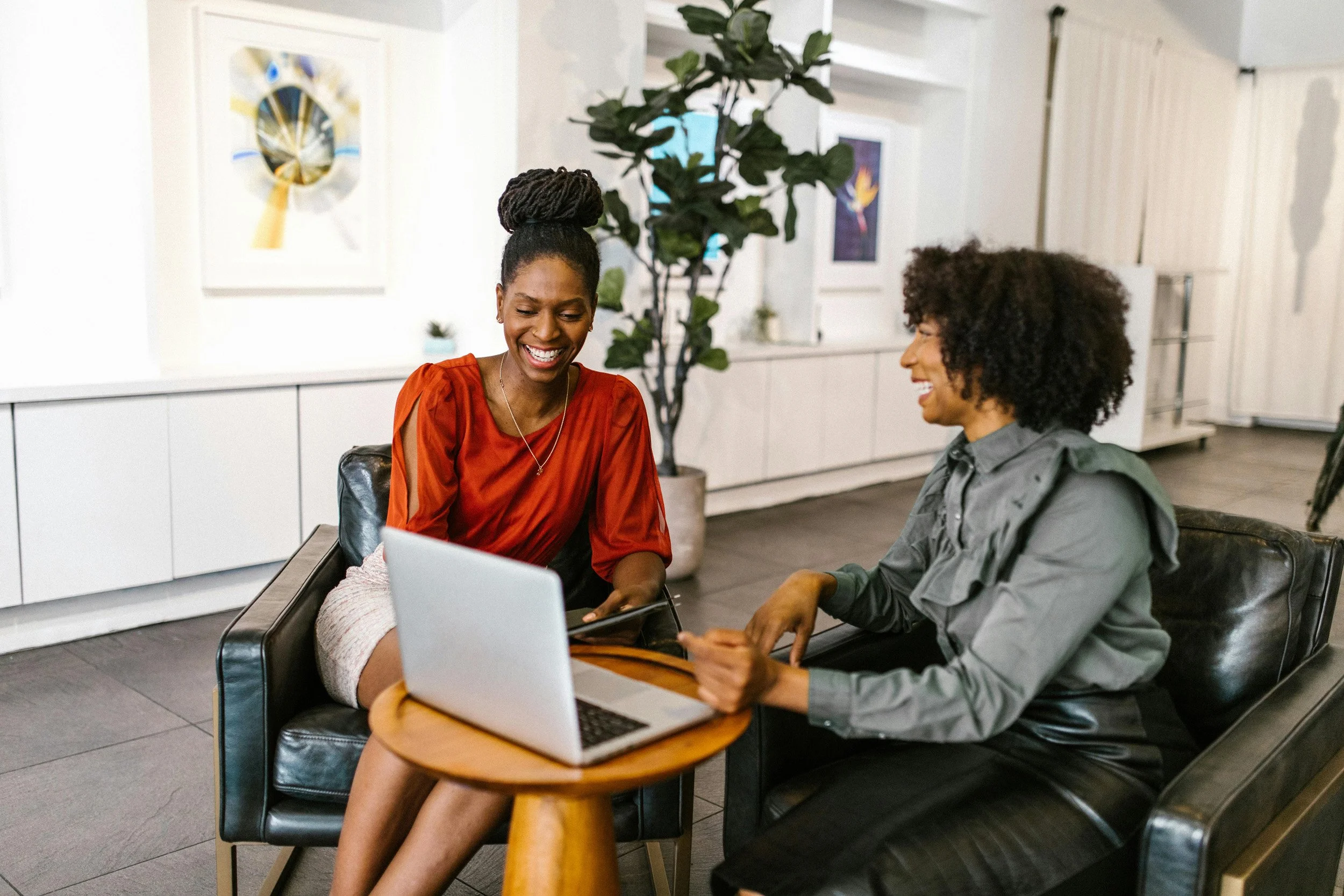 Two women sitting on black leather chairs, looking at a laptop on a small round wooden table between them, smiling and talking in a modern office or lounge with art on the walls and a large plant in the background.