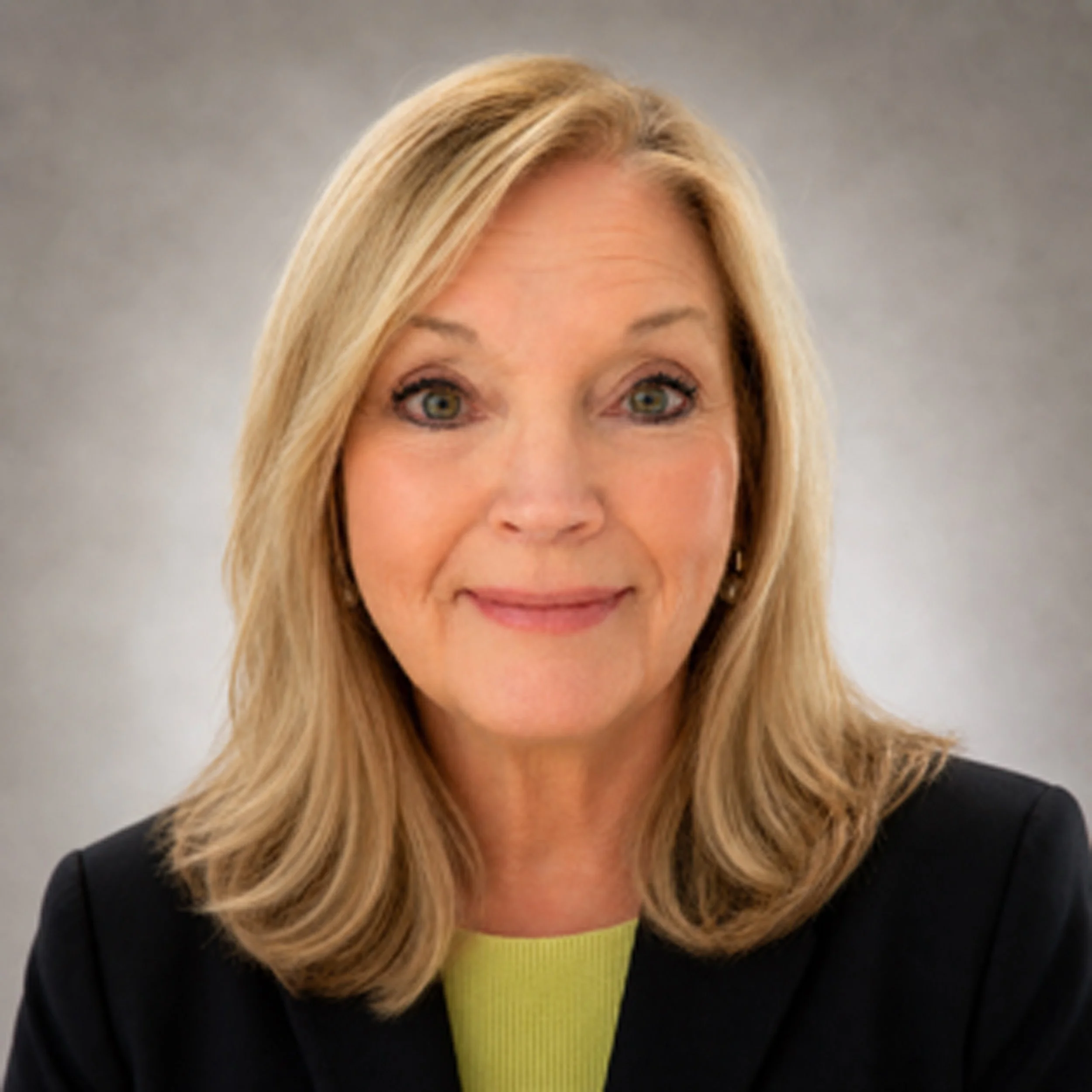 Close-up of a smiling woman with long blonde hair, wearing a dark blazer and a light green shirt, against a plain gray background.