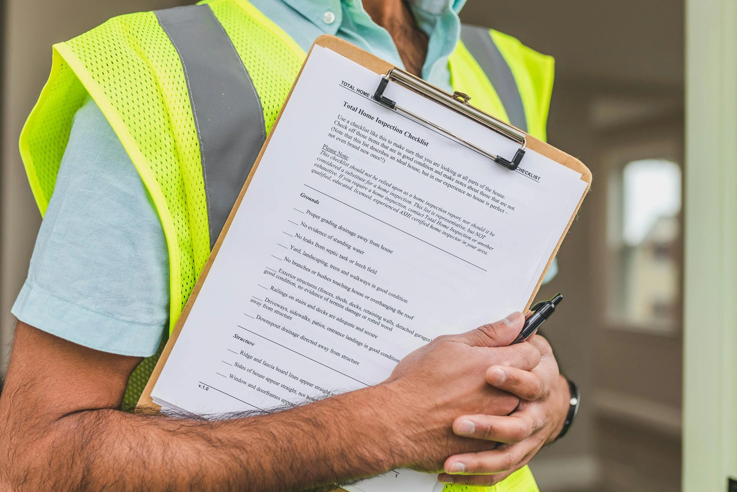 Close-up of a person wearing a high-visibility safety vest and holding a clipboard with a home inspection checklist, with a pen in their hand.