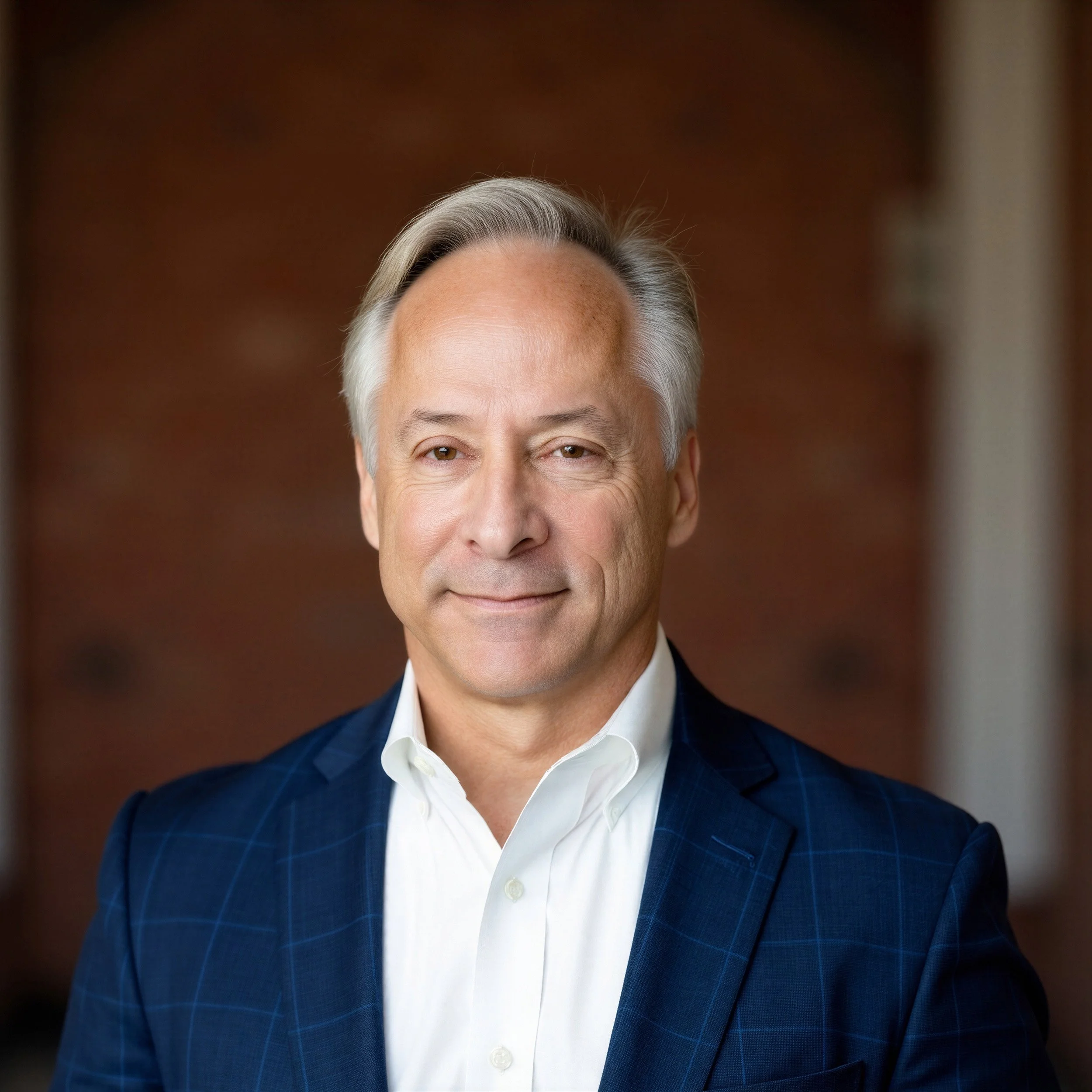 Close-up of a man with gray hair wearing a dark blue plaid suit and white shirt, smiling slightly, against a blurred brown background.