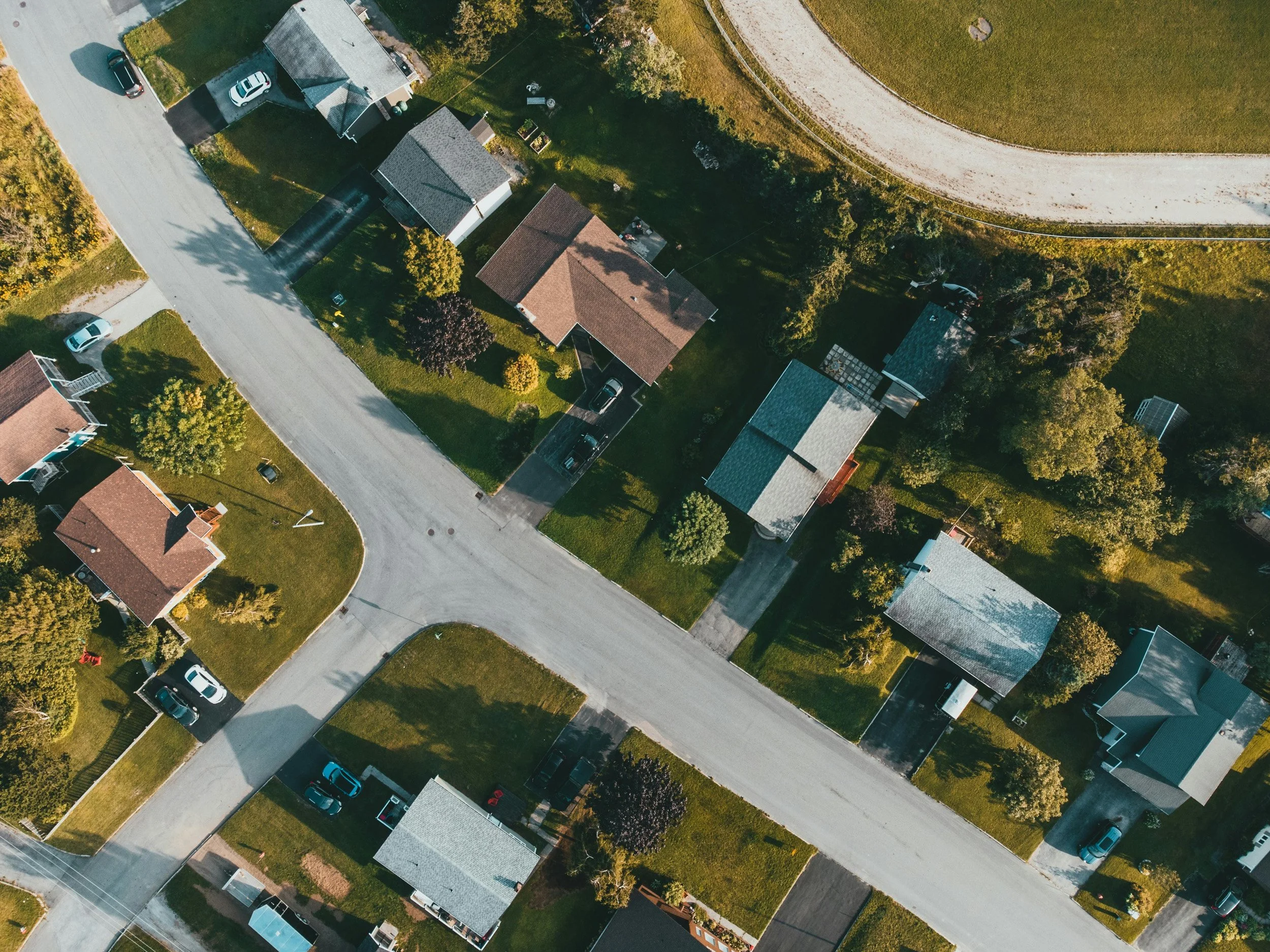 An aerial view of a residential neighborhood with several houses, each with a lawn, trees, and parked cars, along a curved road with a gravel pathway and open green space.