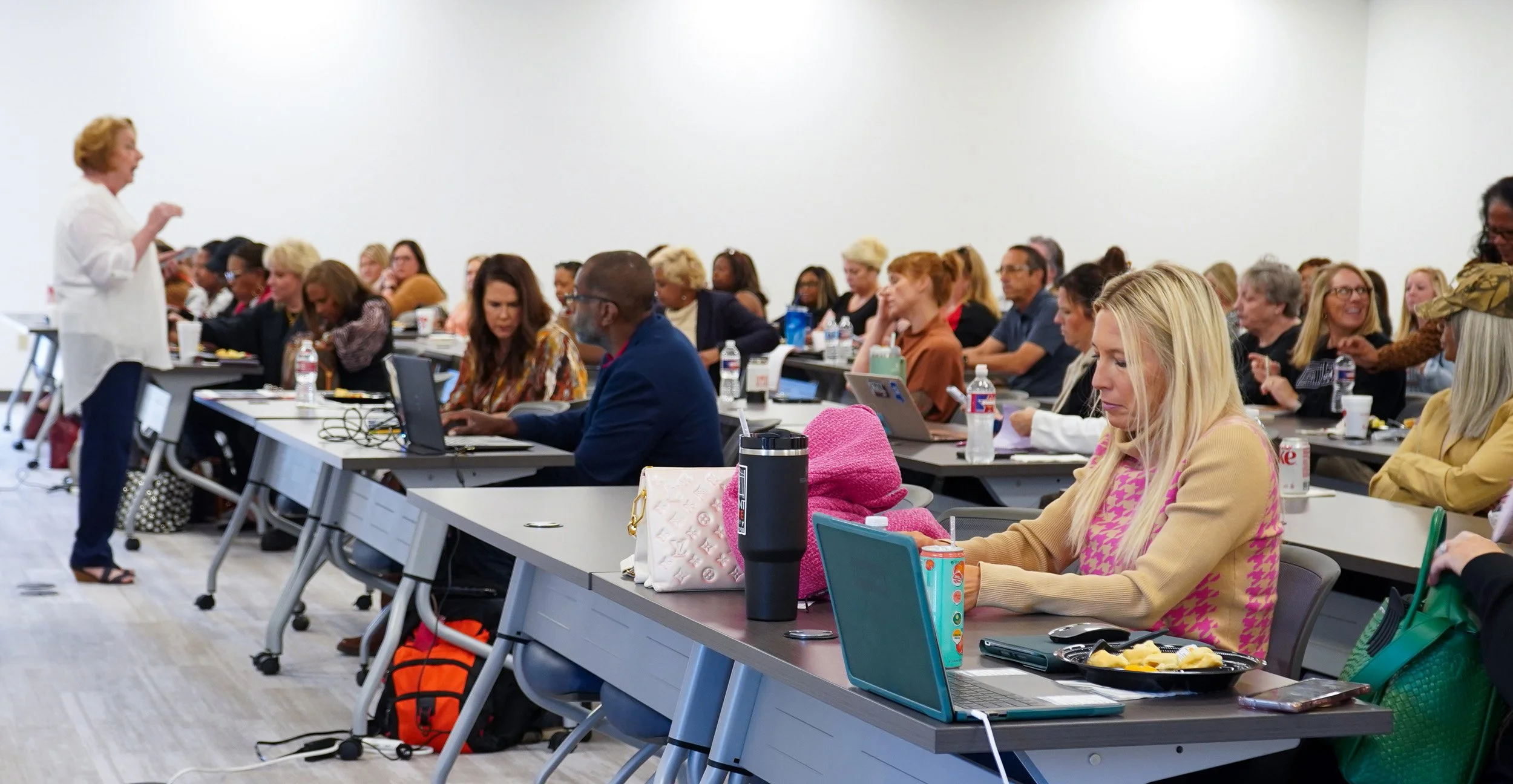 A woman with short red hair speaking in front of a group during a conference or workshop in a large white-walled room. Attendees are seated at long tables with laptops, notebooks, and beverages, listening attentively.