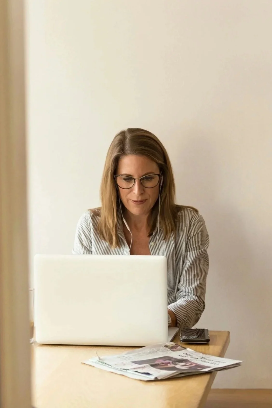A woman with shoulder-length brown hair, glasses, and earphones working on a laptop at a desk, with a newspaper and a smartphone nearby, in a room with plain white walls.