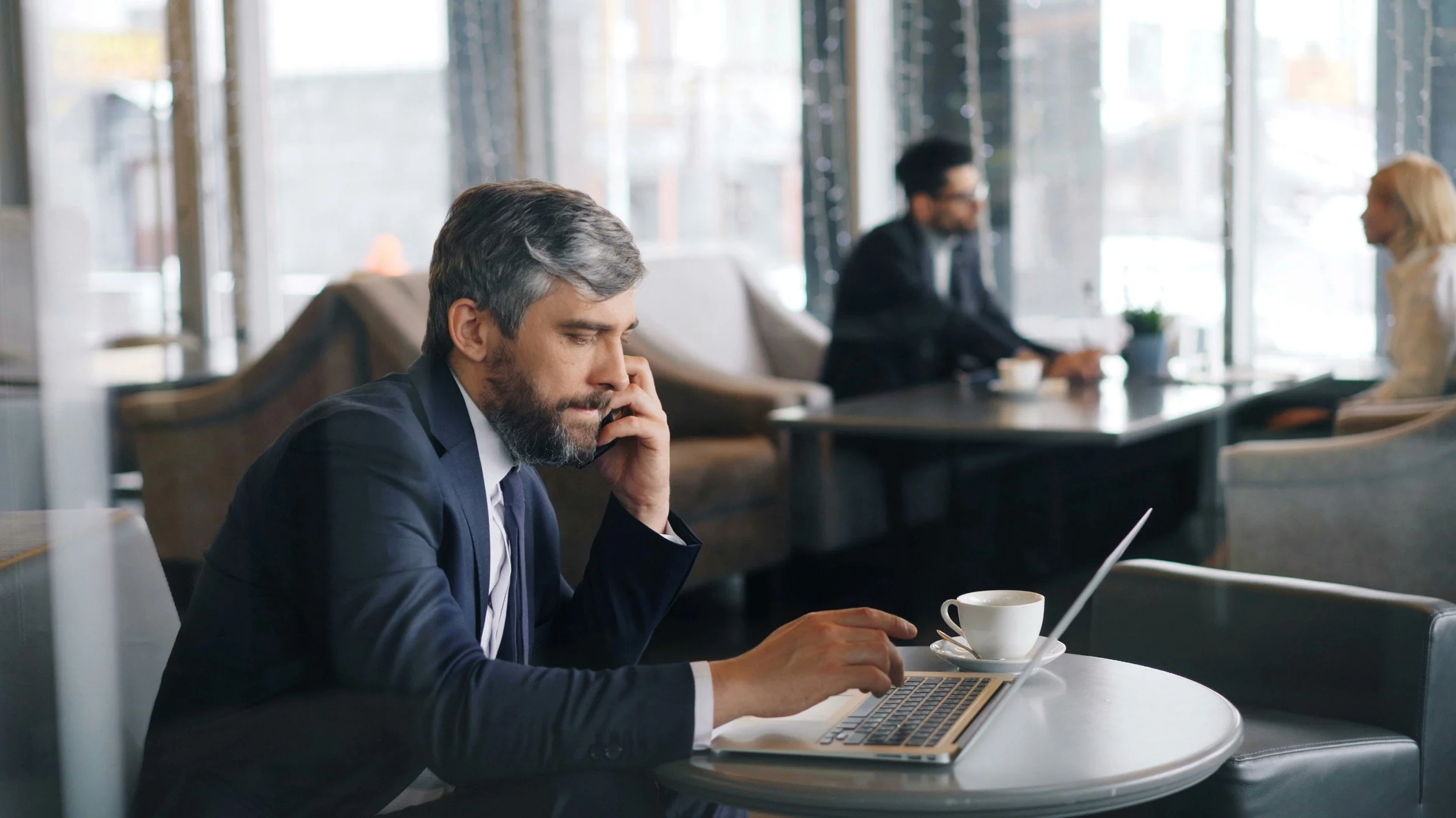A middle-aged man with gray hair and a beard, dressed in a suit, sitting at a table in a coffee shop, working on a laptop and talking on his phone, with a coffee cup nearby.
