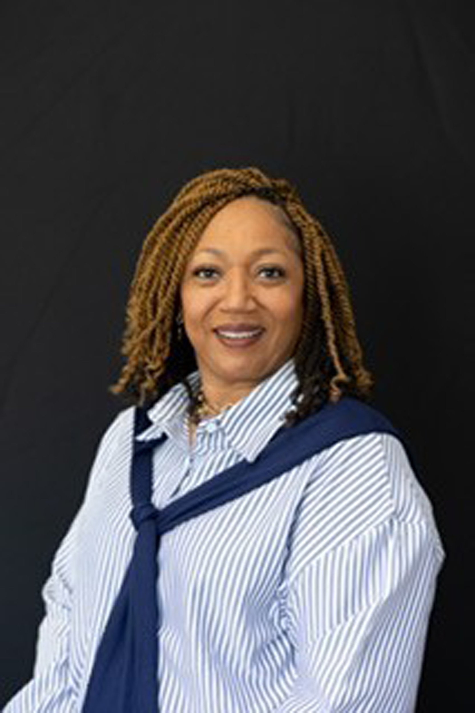 A woman with shoulder-length braided hair smiling in front of a black background, wearing a striped shirt and dark blazer.
