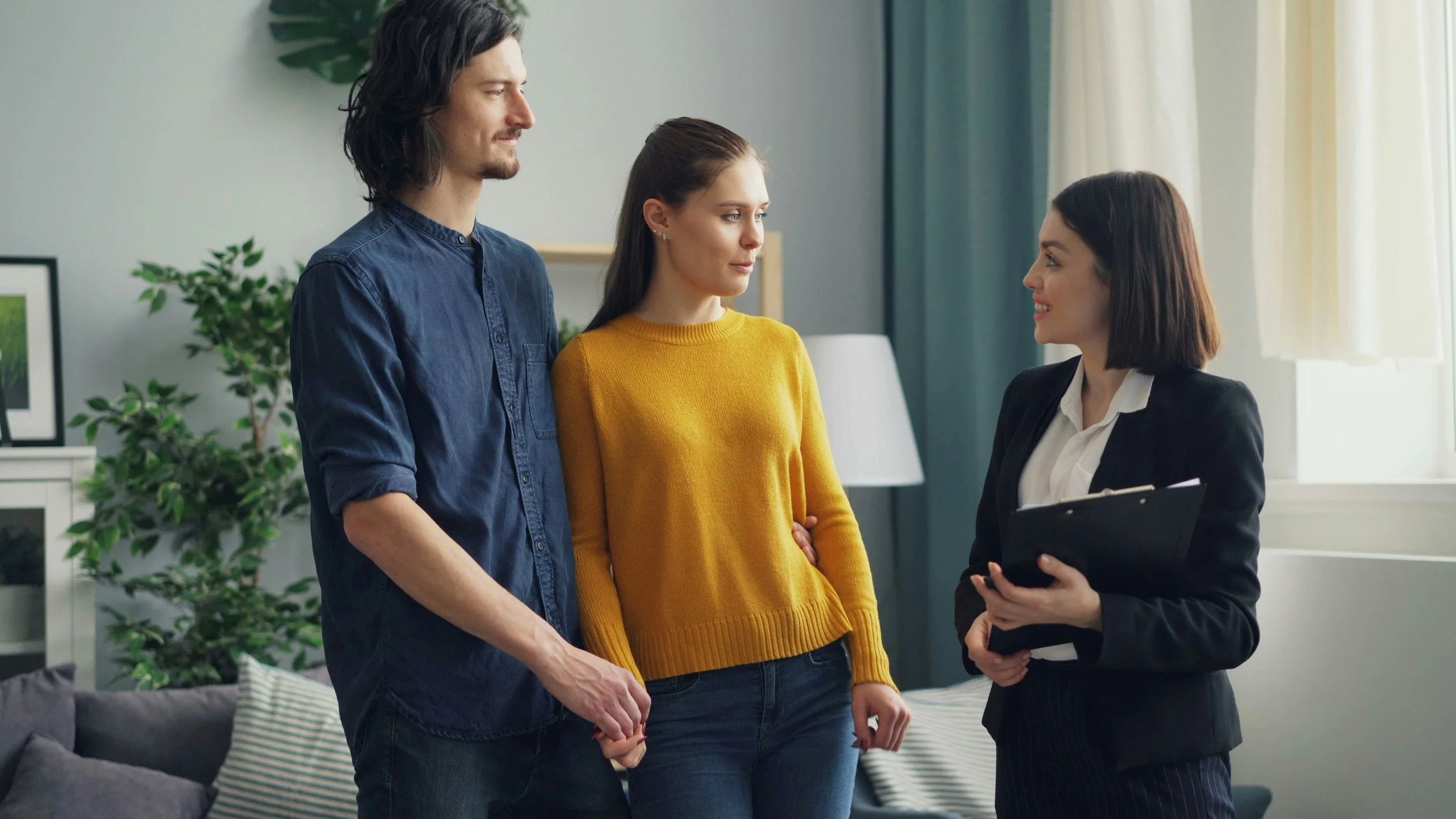A man and woman standing with a woman holding a folder, engaging in a conversation in a living room.