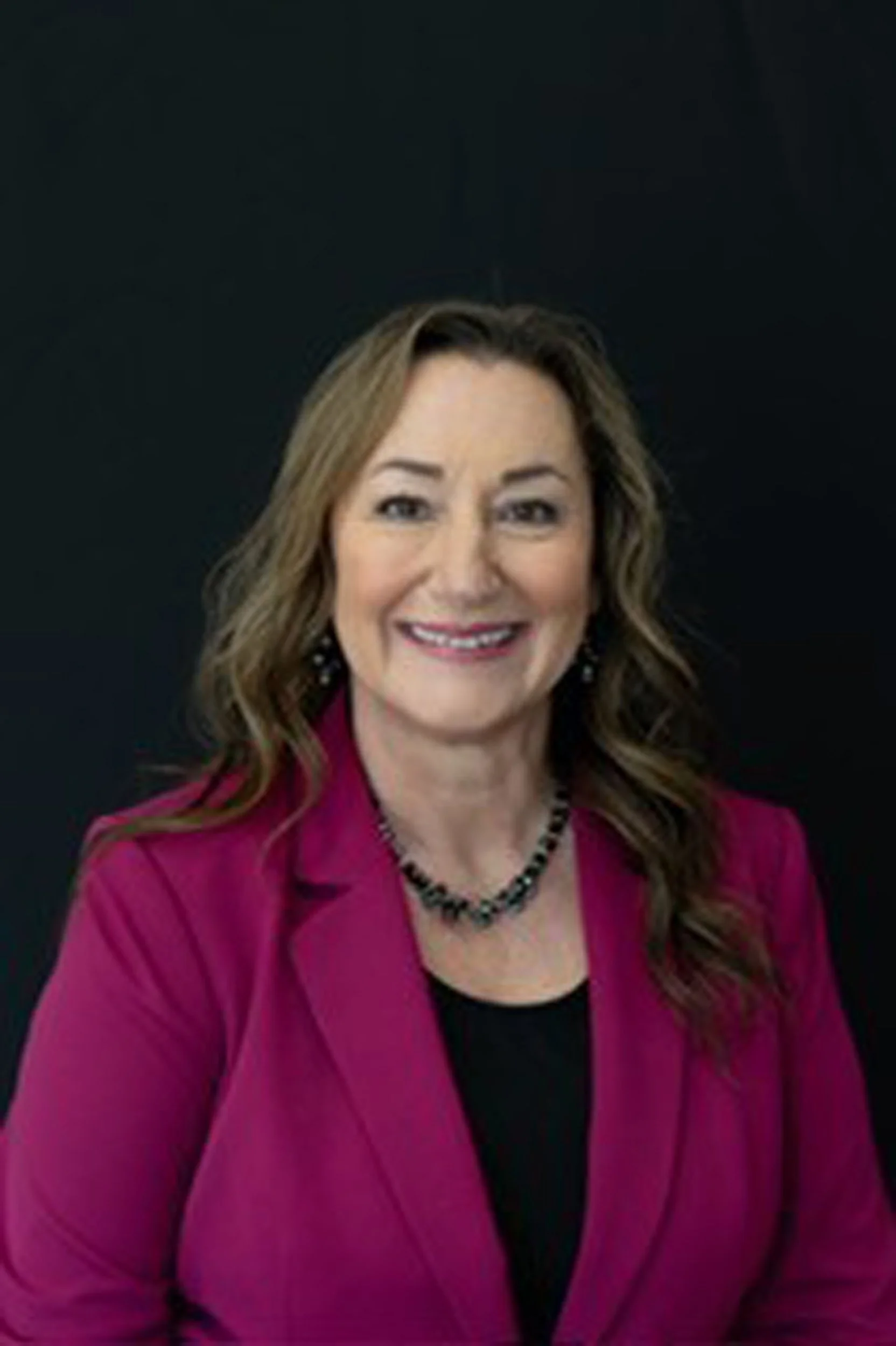 Headshot of a woman with shoulder-length wavy hair, wearing a pink blazer, black top, and a black beaded necklace, smiling against a black background.