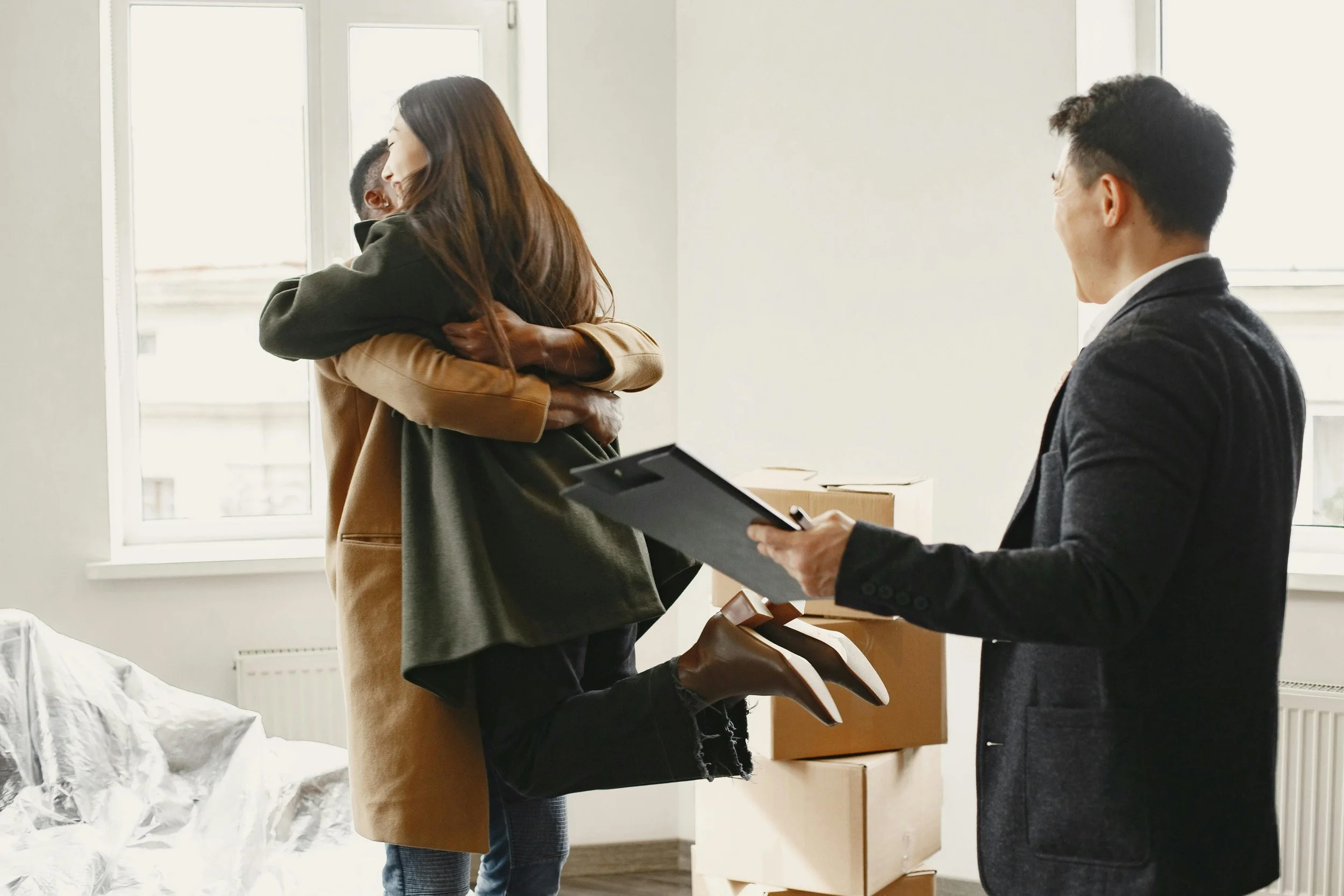 Three people in a room with boxes and a bed. A woman is hugging a little girl, and a man is holding a clipboard while facing them.