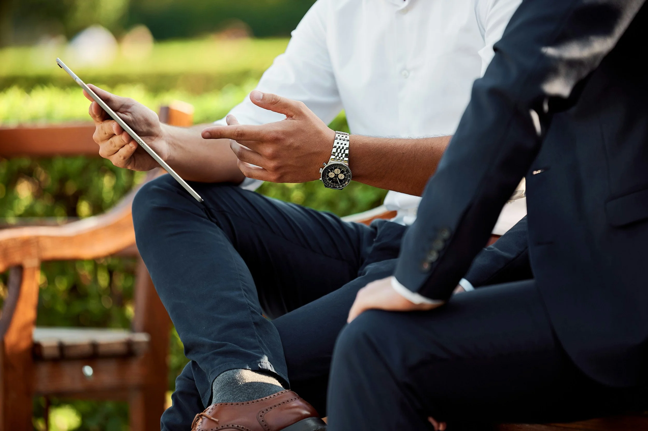 Two men sitting outdoors on a park bench, one is wearing a white shirt and the other is wearing a dark suit, both engaged in a conversation, with one holding a tablet device.