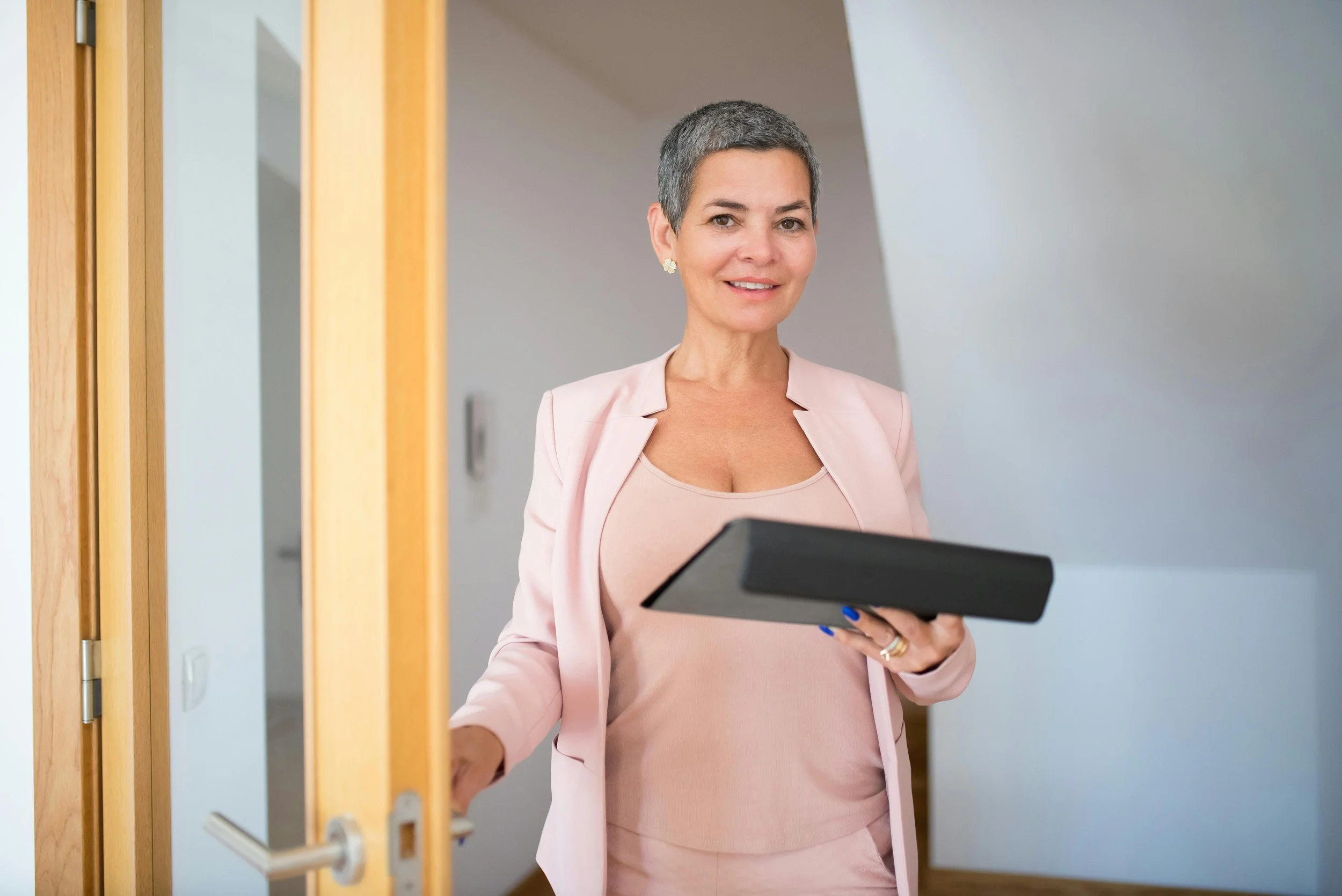 A woman with short gray hair wearing a pink blazer and dress holding a clipboard, standing at a door with a wooden frame.