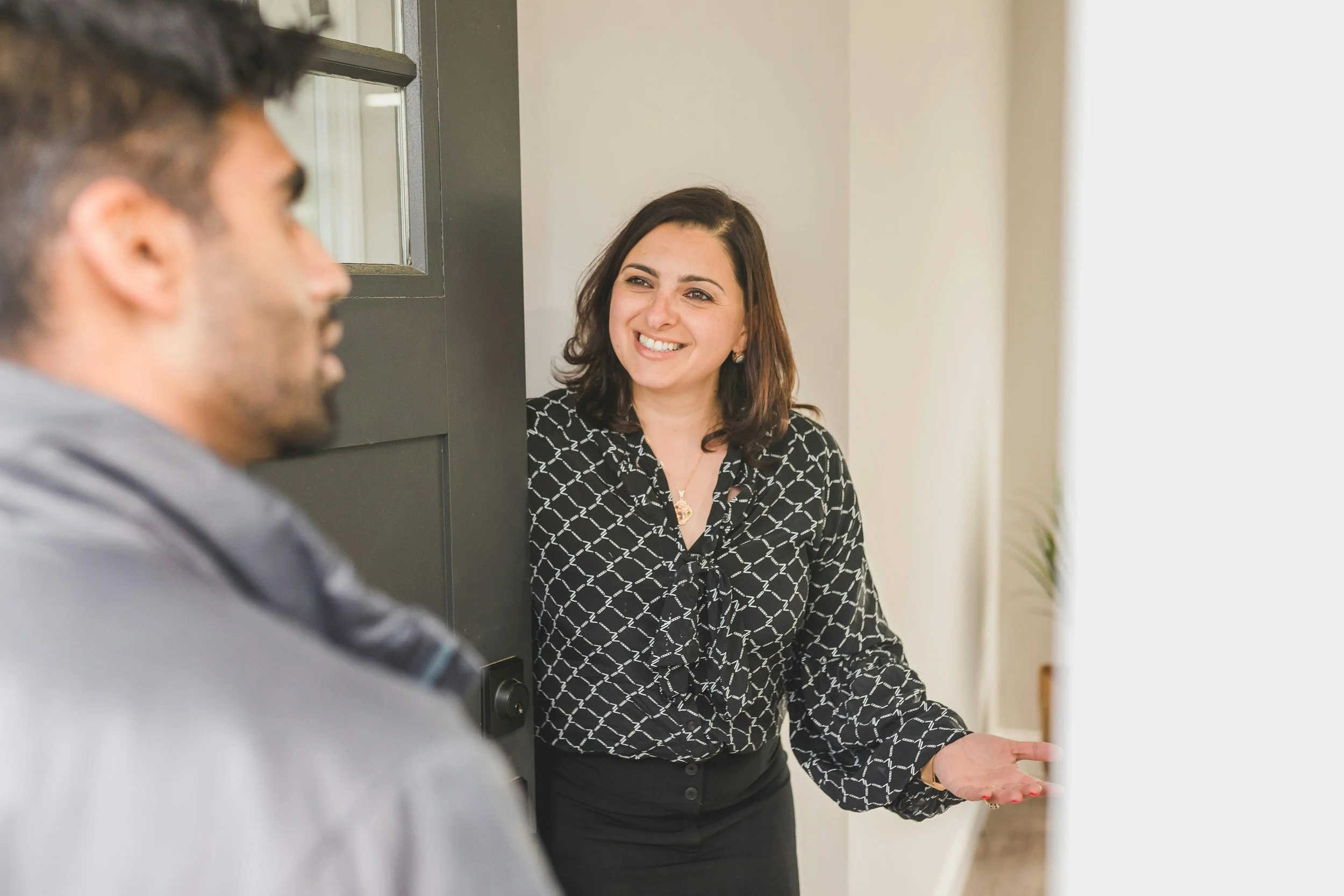 A woman smiling and gesturing with her hands while talking to a man who is out of focus in the foreground. They are indoors, near a door and window.
