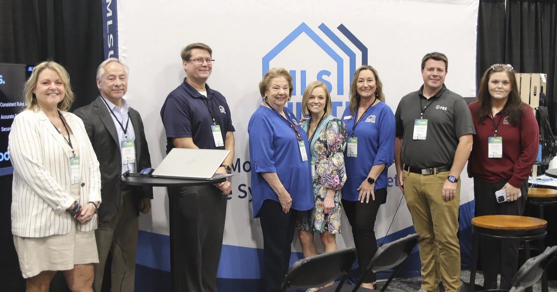 Group of eight people standing in front of a banner that reads 'US' and a house-shaped logo, at an event or conference.