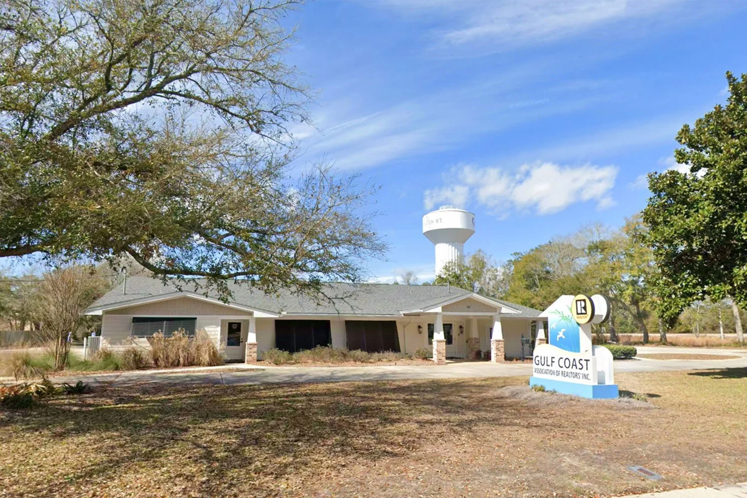 A small, single-story building with a gray roof and light-colored walls, surrounded by trees and a parking area, with a water tower visible behind the building. There is a sign that reads "Gulf Coast Association of Realtors Inc." in front of the building.