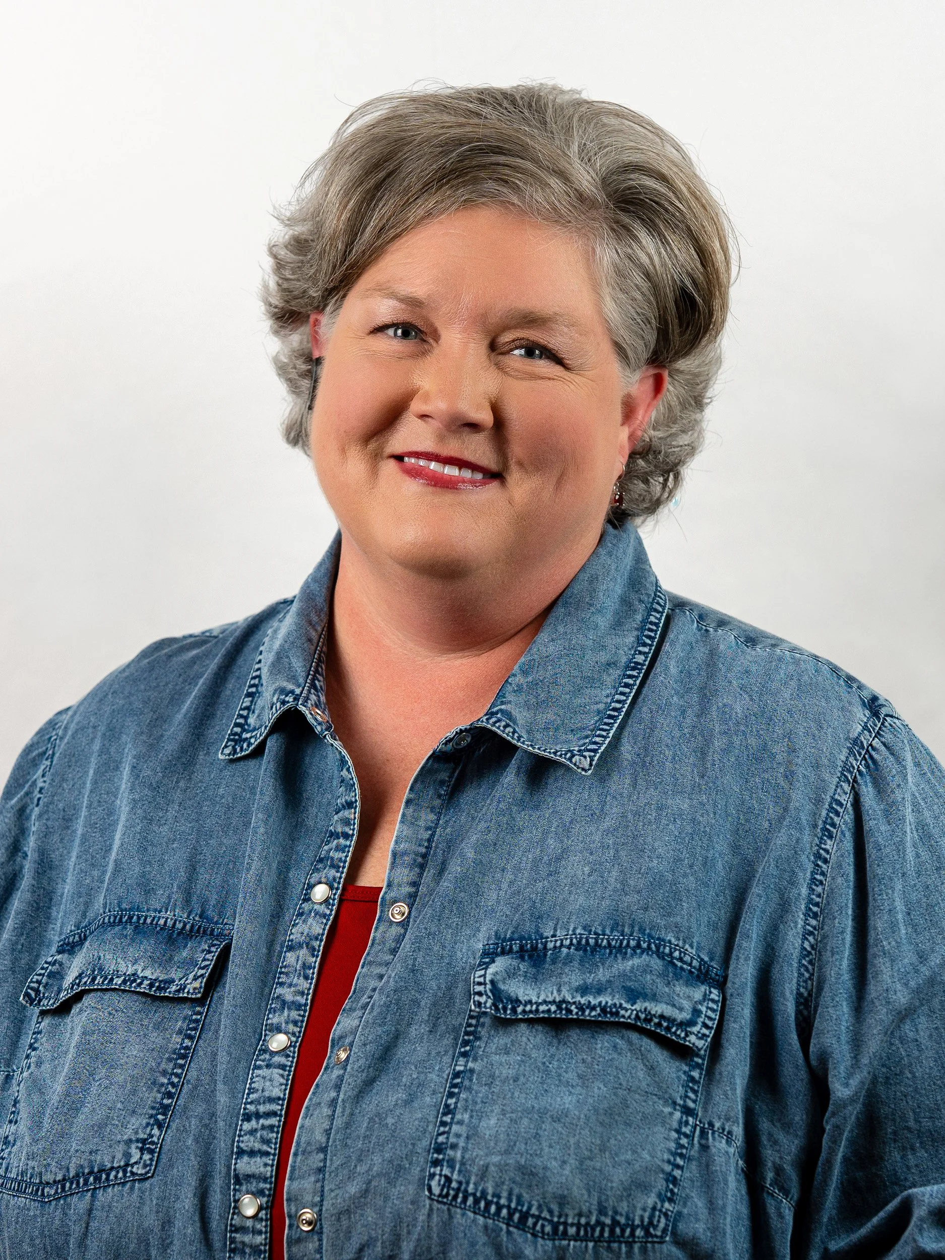 A woman with short gray hair smiling, wearing a blue denim jacket and a red top inside.