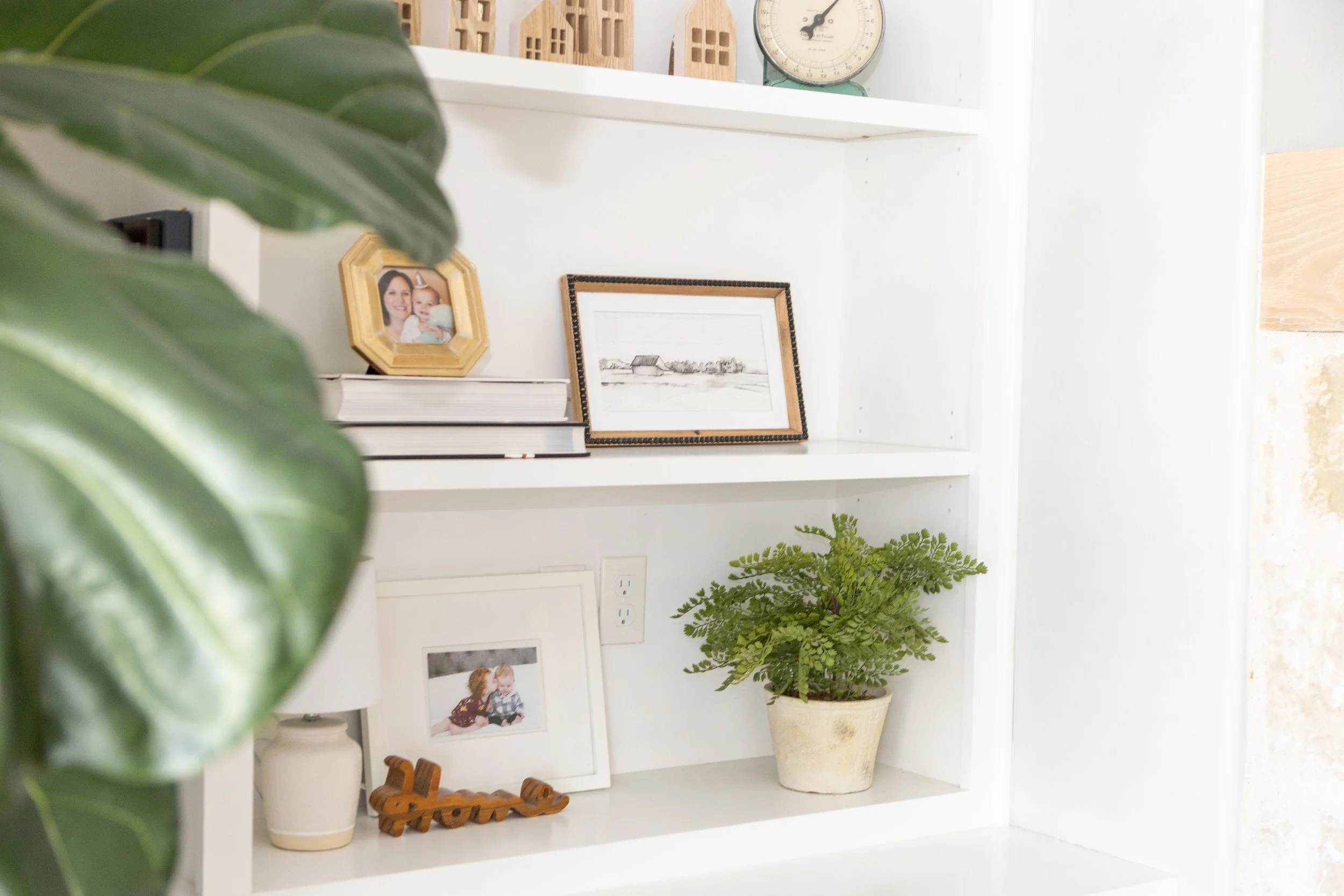 White bookshelf with decorative items including a green potted plant, framed photographs, small wooden houses, a vintage clock, and a black-framed drawing of a barn landscape.