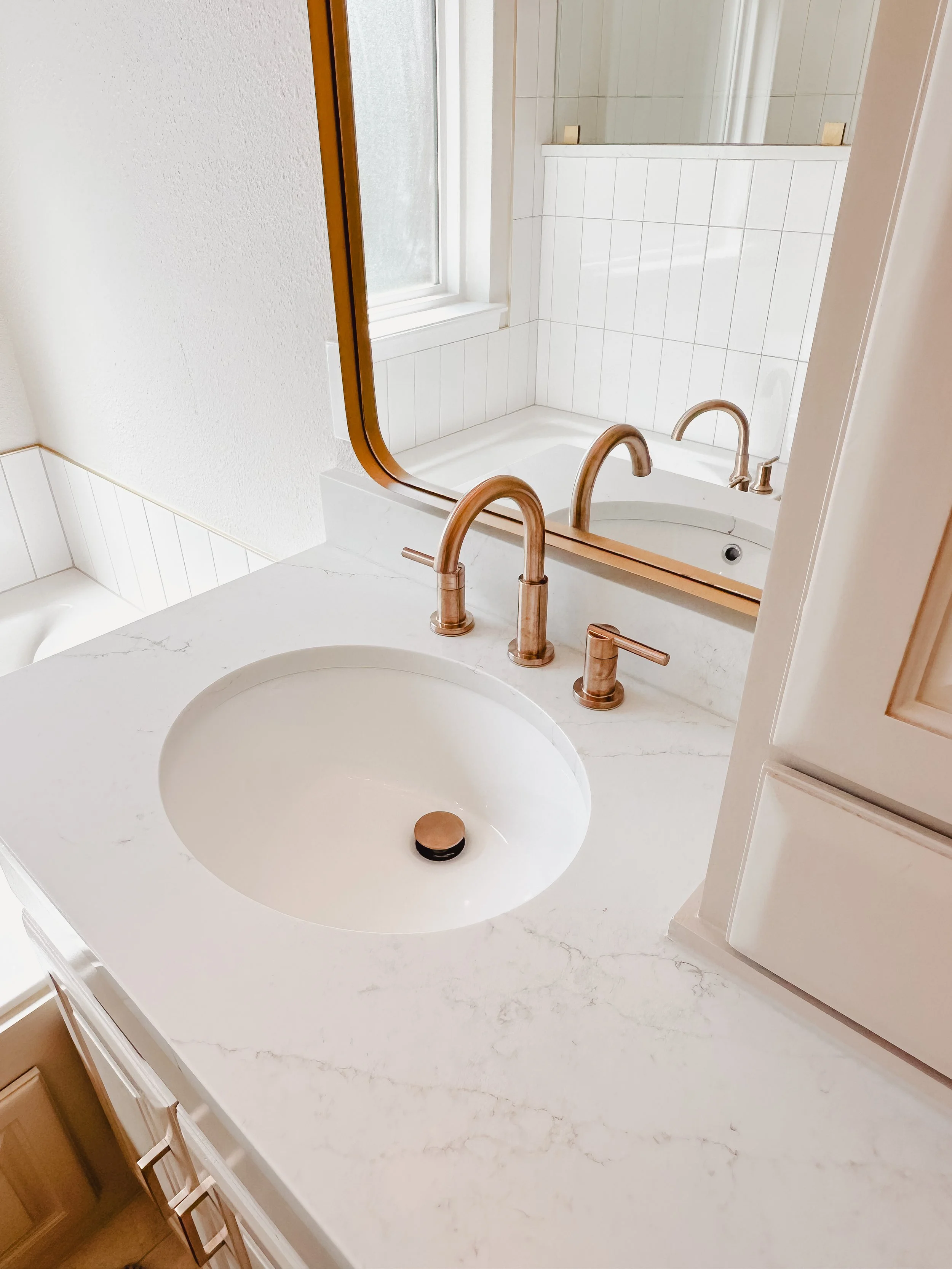 Bathroom sink with a marble countertop, a round white basin, brushed copper faucets, a large mirror, and a window with frosted glass.