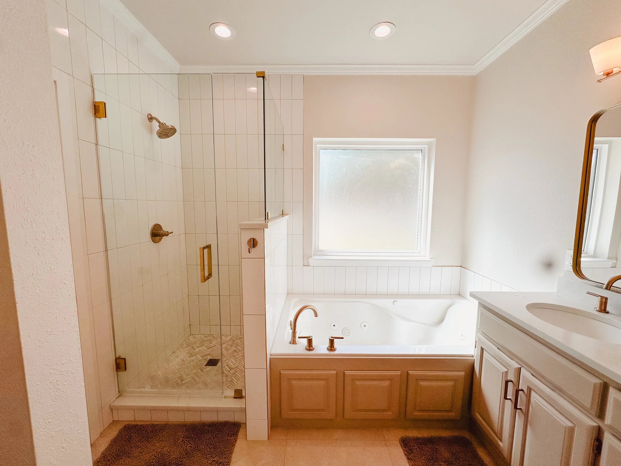 A bathroom with a shower area enclosed with glass, white tiled walls, beige floor tiles, and a window allowing natural light.