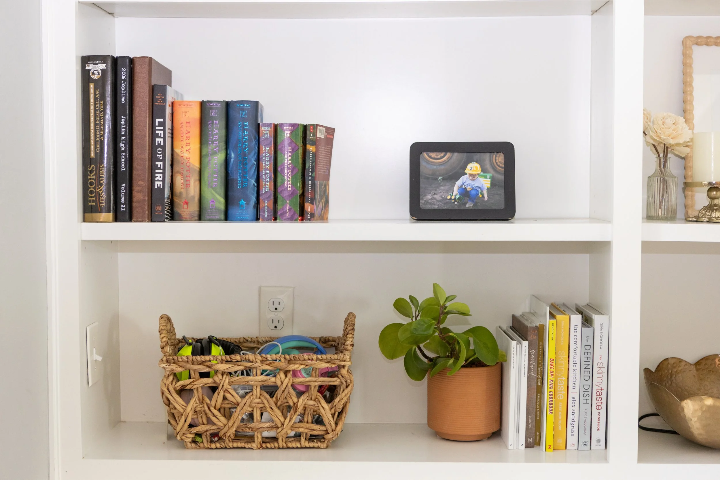 White bookshelf containing a row of books, a digital photo frame displaying a child playing, a potted plant, and a vase with flowers.