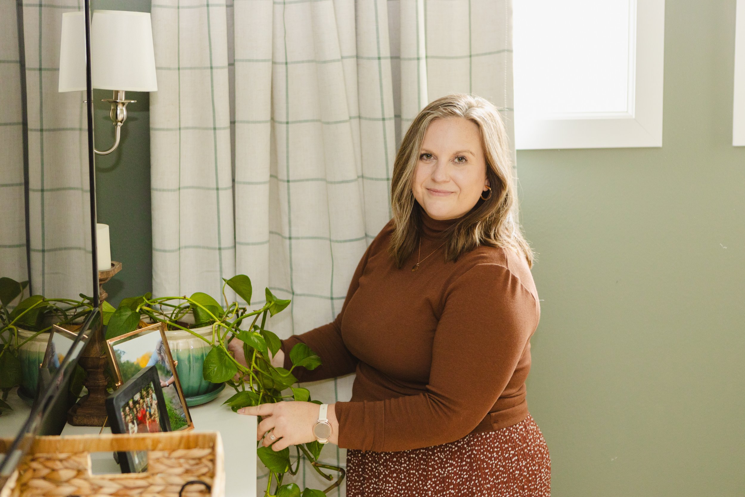 A woman with shoulder-length light brown hair, wearing a brown long-sleeve top and a red patterned skirt, standing indoors near a table with framed photos and a potted plant. She is smiling and pointing at one of the photo frames.