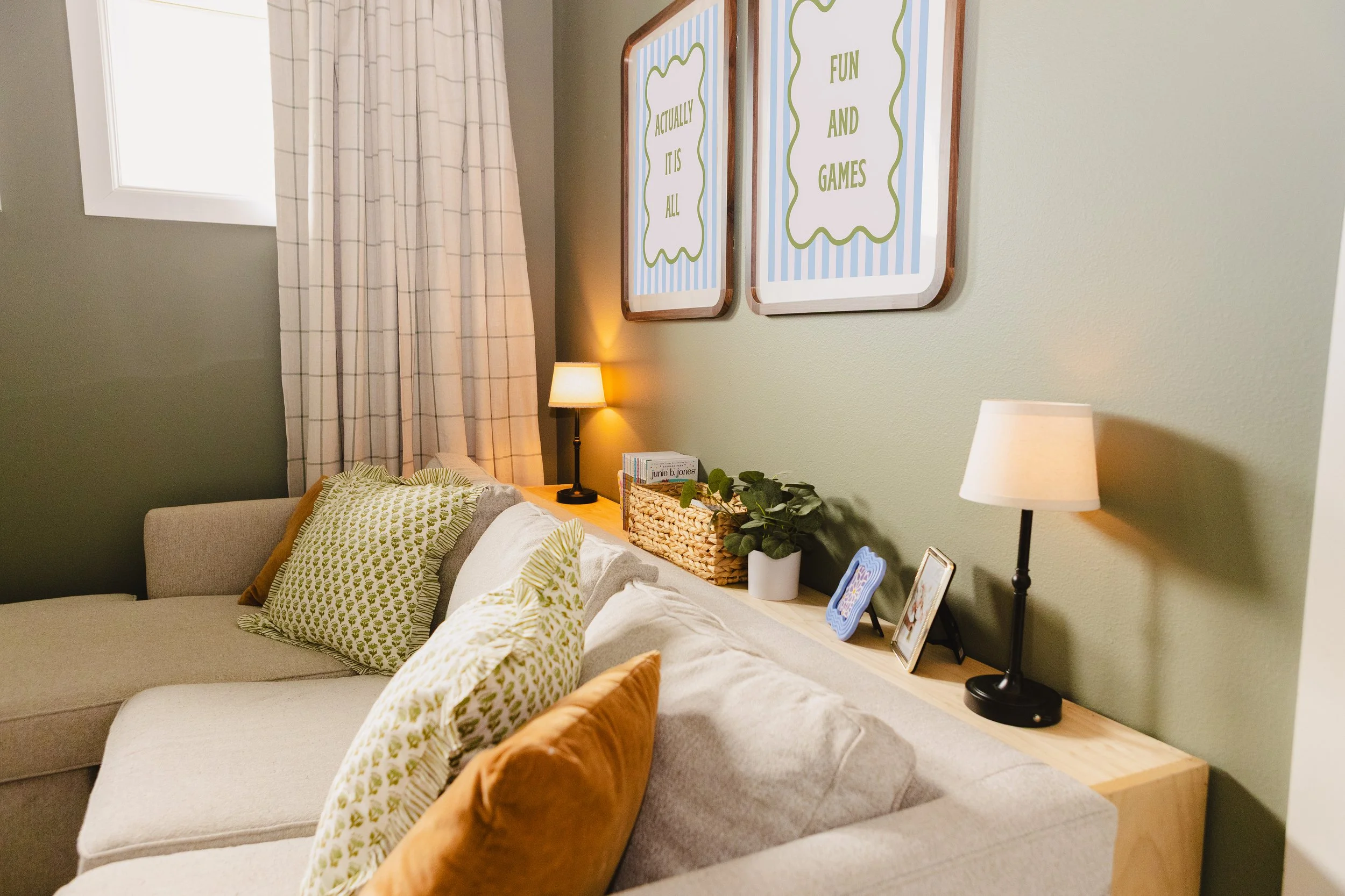 Living room with a beige sofa adorned with green and rust-colored pillows, a wooden side table with framed photos, a basket with green plants, and a lamp on a narrow wooden shelf. The wall behind has two framed art pieces with blue and white striped 