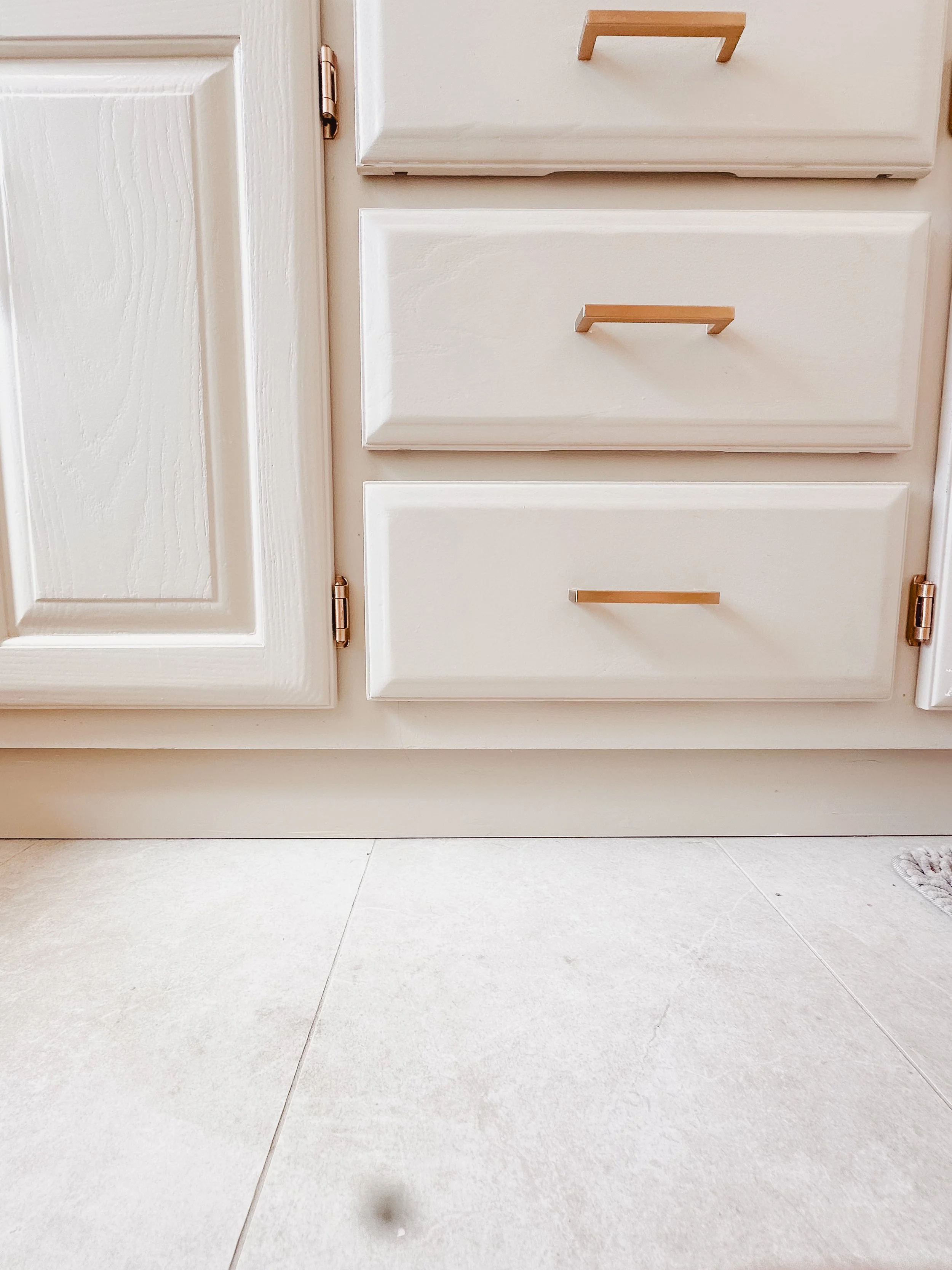 Close-up of white kitchen cabinets with wooden handles and a beige countertop, with light-colored tiled floor beneath.
