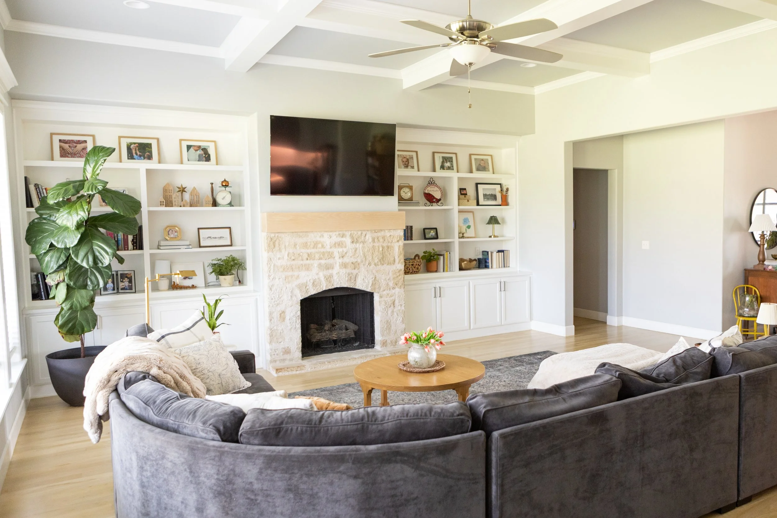 Living room with a large L-shaped gray velvet sofa, a round wooden coffee table with a flowers in a vase, built-in white shelves with framed photos and decorations, a stone fireplace, a large flat-screen TV, a ceiling fan, a potted plant, and a stand
