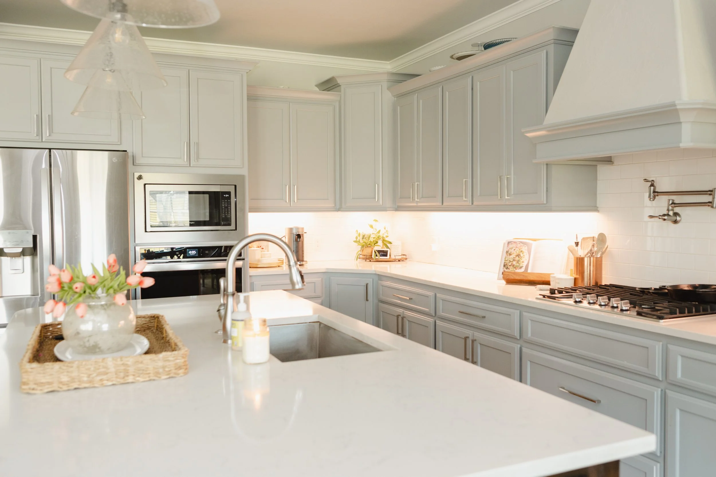 A modern kitchen with white cabinets, stainless steel refrigerator, microwave, and gas stove. Decor includes a flower arrangement, a small plant, and kitchen utensils on a white countertop.