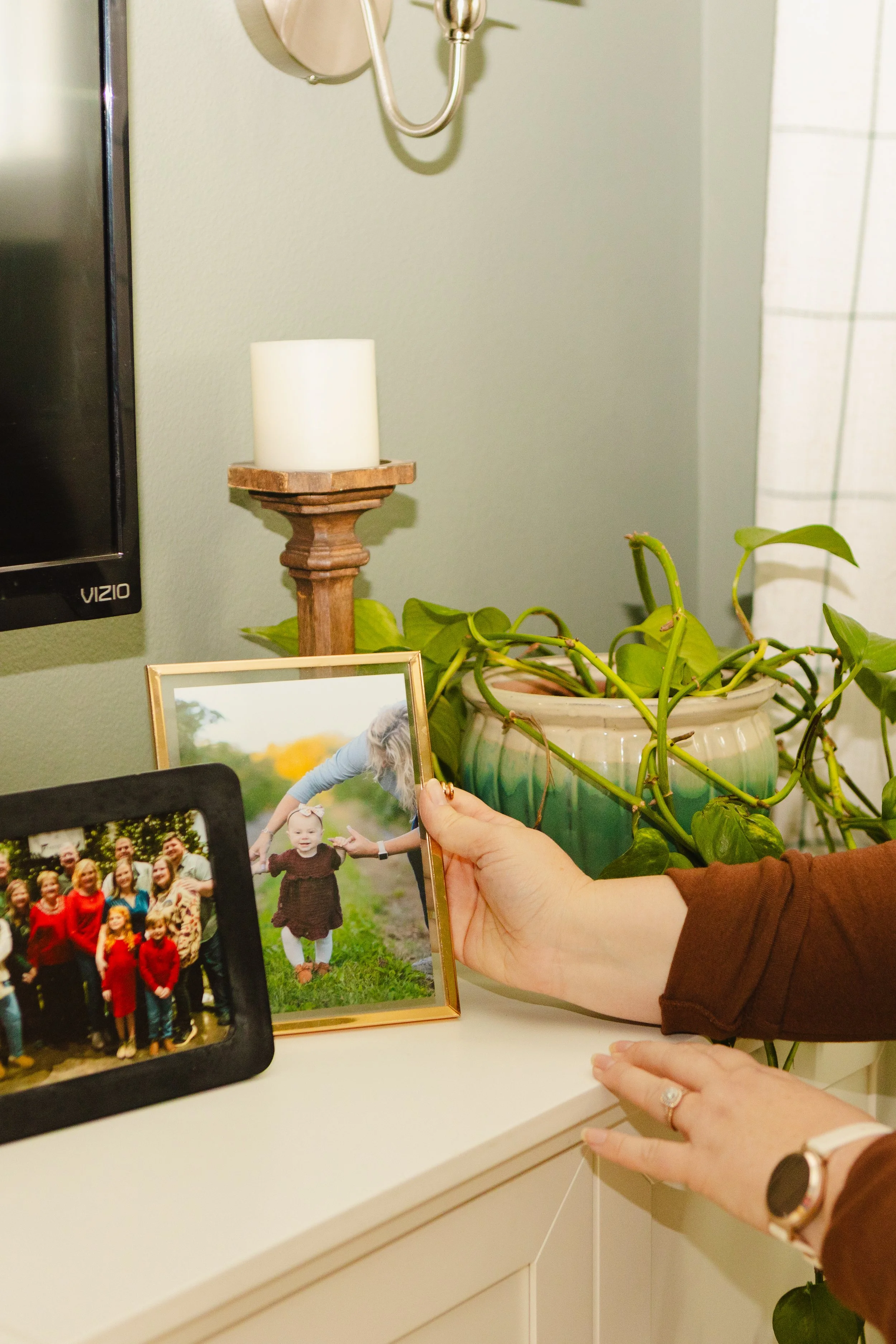 A person with a ring on their finger is holding a picture of children standing outdoors, with a framed photo of a family group on the desk. Behind the desk is a houseplant in a ceramic pot, a small wooden candle holder with a white candle, and a wall-mounted lamp.
